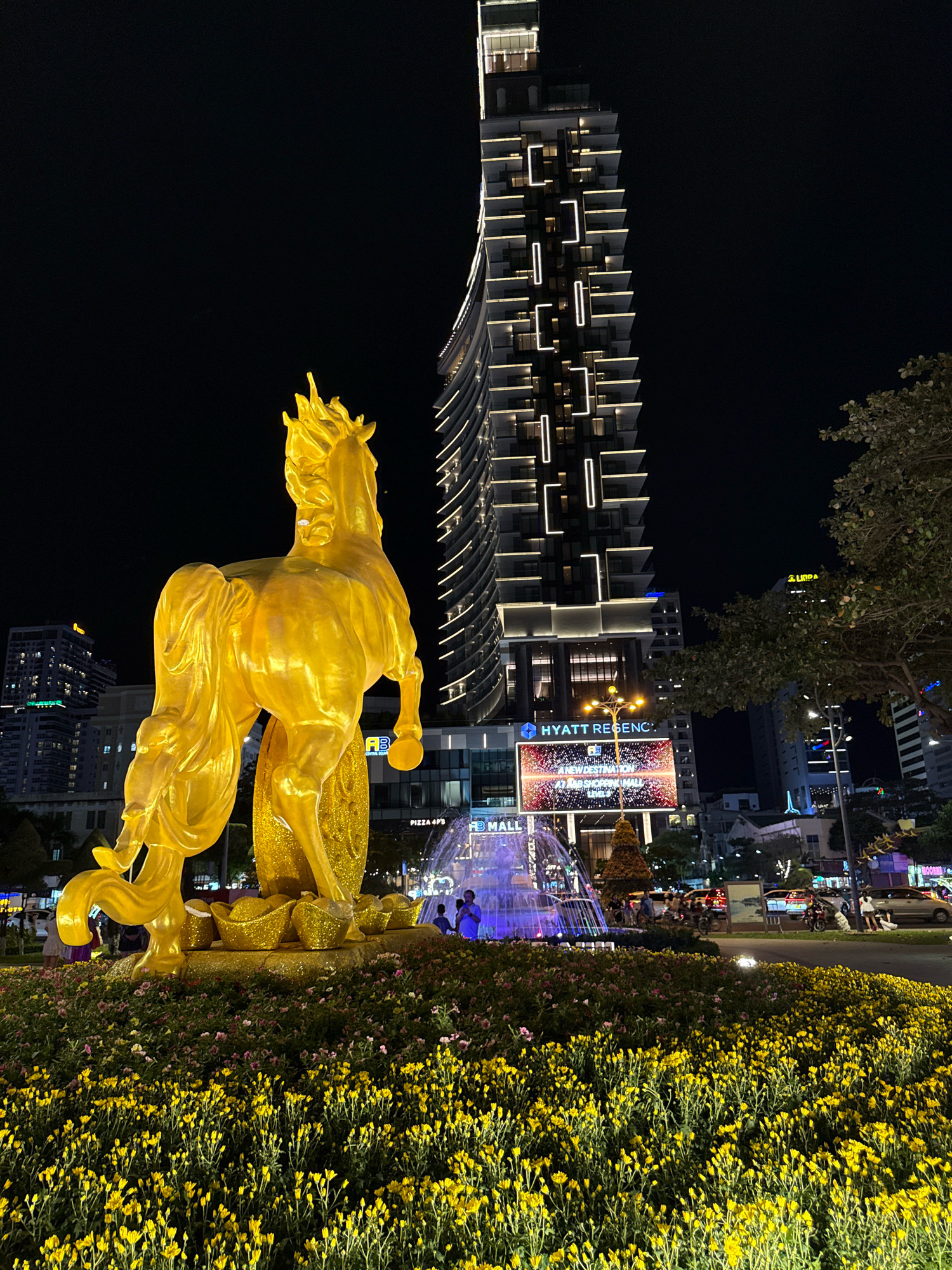 This is a nighttime photograph taken in Nha Trang, Vietnam, showcasing a large, ornate golden horse statue positioned in the foreground. The statue is surrounded by a bed of vibrant yellow flowers. Behind the statue, a modern, illuminated skyscraper with a distinctive architectural design pierces the dark sky. At the base of the skyscraper, a large LED screen displays an advertisement for "Hyatt Regency" and "A New Destination at AB Shopping Mall." A multi-tiered fountain with illuminated blue water jets is situated in front of the screen. Several individuals, appearing as silhouettes, are visible near the fountain and on a nearby street, where traffic, including cars and motorcycles, is present. Streetlights and the lights from other buildings contribute to the illuminated urban environment.