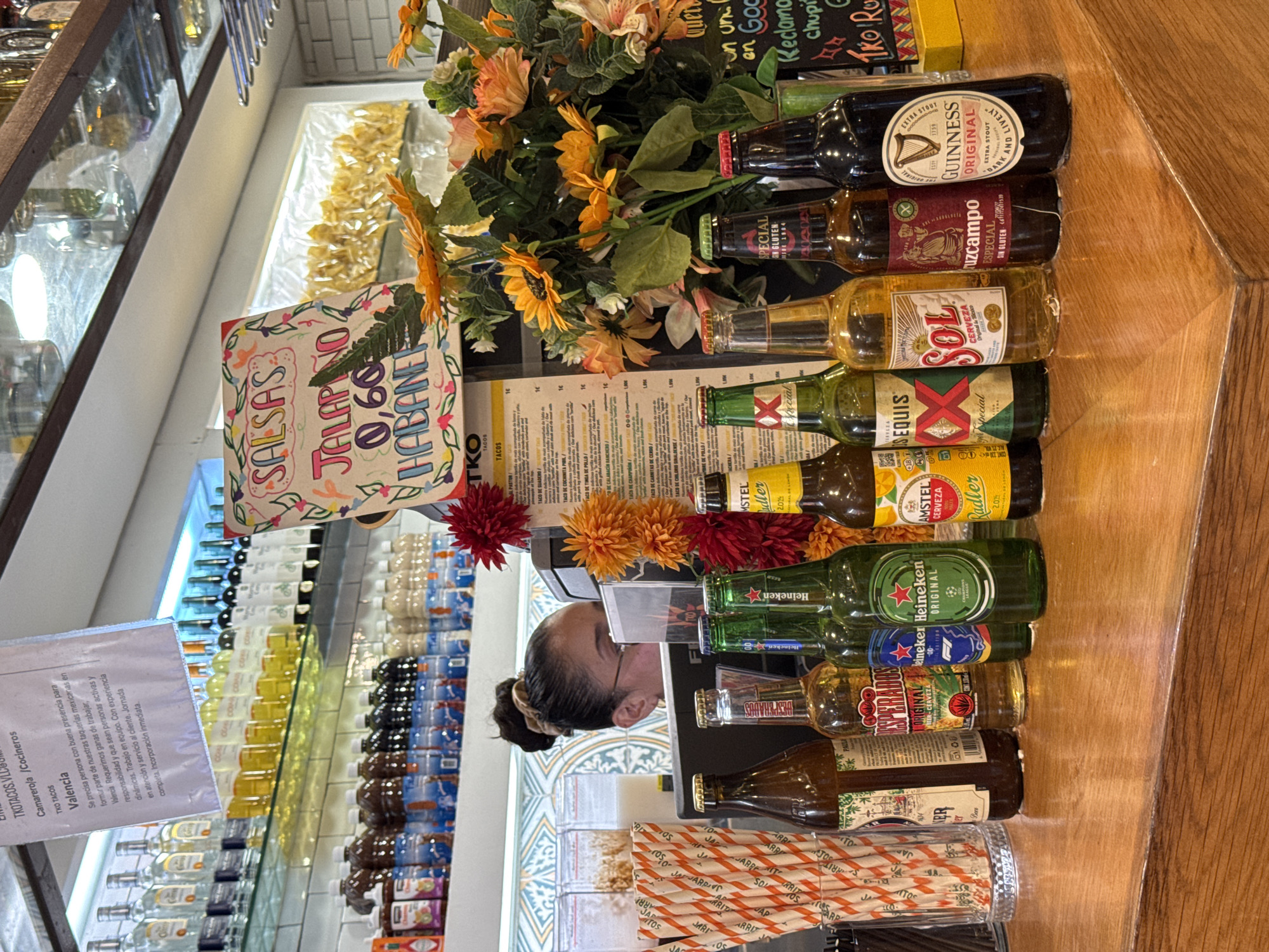 The image depicts a bar counter in Valencia, Spain, with various bottles of beer and other beverages displayed. In the background, shelves are stocked with more bottles, including spirits and wine. A sign with handwritten text about salsas and prices is visible, as well as a menu board. A woman wearing glasses is partially visible behind the counter, appearing to be working. Decorative flowers, predominantly orange and yellow sunflowers, are arranged on the counter. The lighting suggests an indoor setting, likely a restaurant or bar.
