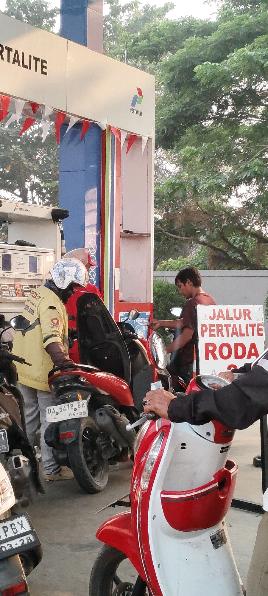 A street-level, eye-level shot captured outdoors at a Pertamina gas station in Banjarbaru, Indonesia, features several motorbikes and individuals. The Pertamina brand signage is prominent on a white structure above. To the left, a person in a yellow jacket and white helmet is attending to a red motorbike. In the center background, a man in a reddish-brown shirt is interacting with a gas pump. A white sign with red lettering that reads "JALUR PERTALITE RODA" (Pertamina Fuel Lane for Wheels) is visible on the right. The environment appears to be a typical urban setting with trees in the background.