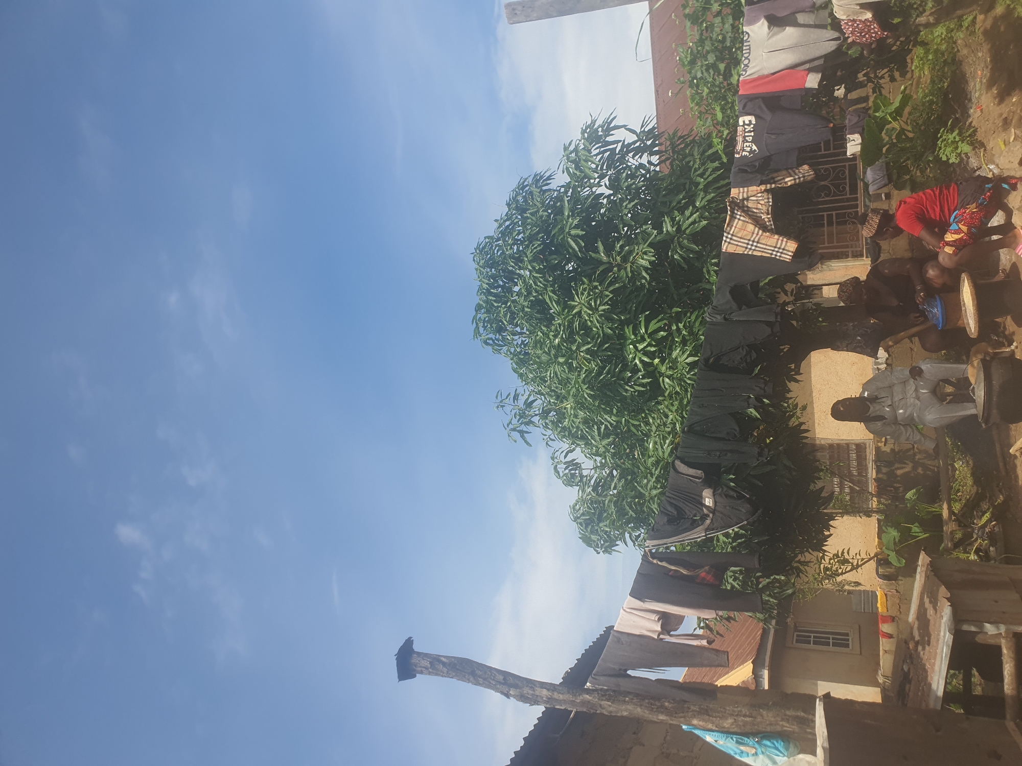 The image depicts an outdoor scene in Bukuru, Nigeria, under a clear blue sky with a few wispy clouds. The orientation of the image is rotated 90 degrees counter-clockwise. In the foreground, a wooden structure, possibly a fence post or part of a roof, is visible. Beyond this, laundry hangs on a clothesline, with various garments including shirts and pants displayed. A lush green tree with dense foliage is prominent in the middle ground.
To the right of the tree, a building with an orange-red brick facade is partially visible. Several people are present in this area. One person, wearing a red shirt and a patterned fabric around their waist, is seated. Another person, dressed in white, is also seated. There are some wooden barrels or large containers in the immediate vicinity. The scene suggests a domestic setting, likely a residential area.
Some text is visible on the clothing hanging on the line. One item has "EXTREME" printed on it. Another garment appears to have a plaid pattern. The overall atmosphere appears to be casual and everyday. The lighting suggests it is daytime, likely mid-morning or afternoon given the brightness and the shadows.