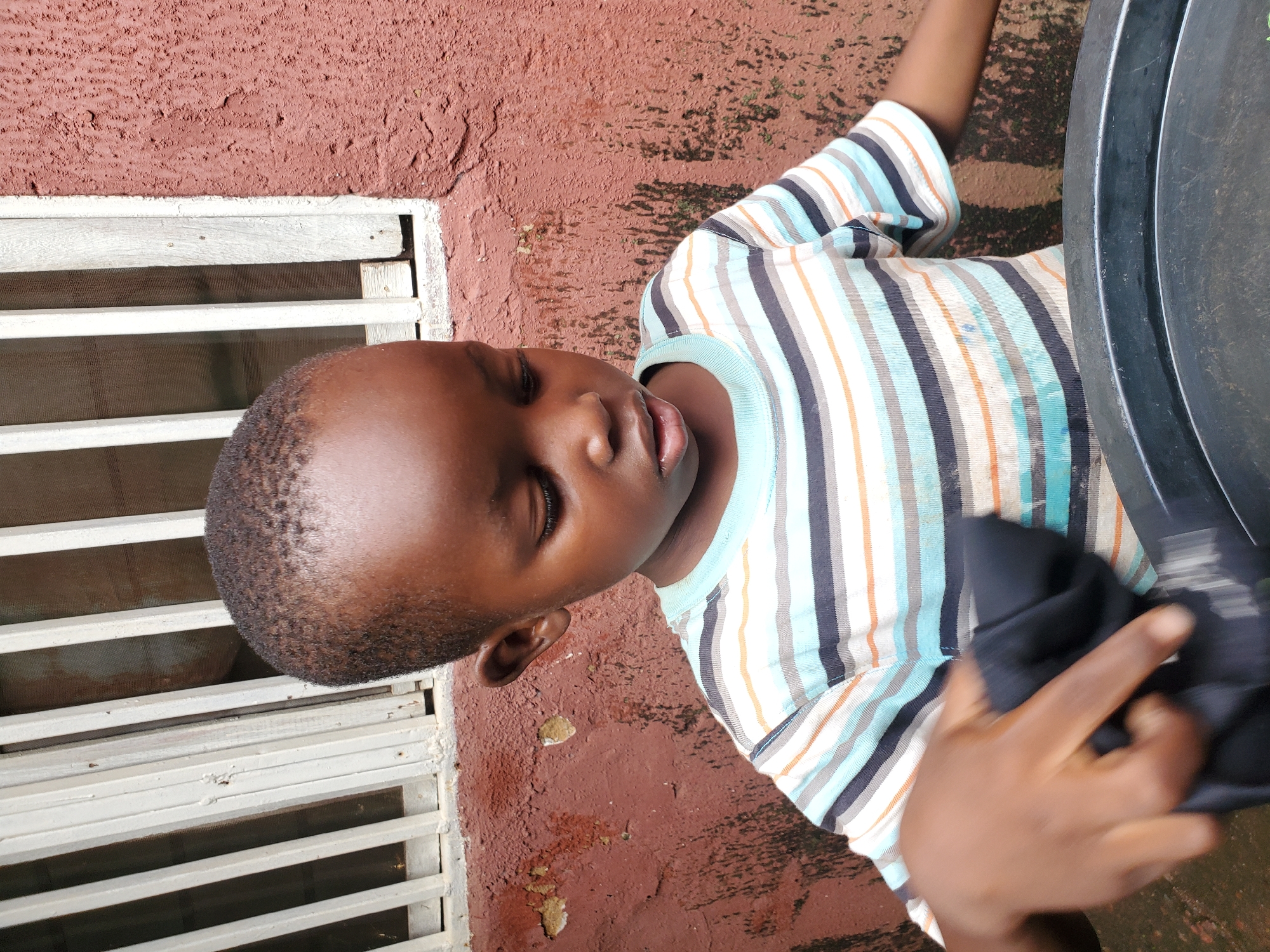 This media file shows a close-up of a young Black child. The child is wearing a striped t-shirt with colors including blue, white, orange, and black. The child's eyes are closed, and their head is tilted back, giving an impression of rest or contemplation. The child's right hand is visible, holding a piece of dark fabric.
The background appears to be an outdoor setting with a textured, reddish-brown wall. To the left, a white window grate is visible. The ground is a similar reddish-brown color with some darker patches, possibly indicating wetness or dirt. The lighting suggests it is daytime, with sunlight casting shadows. No specific location cues are immediately apparent, but the overall scene is casual and domestic.
There is no visible text in the image. The child's expression is neutral, or perhaps peaceful. The focus is on the child and their immediate surroundings. The composition suggests an informal, candid moment.