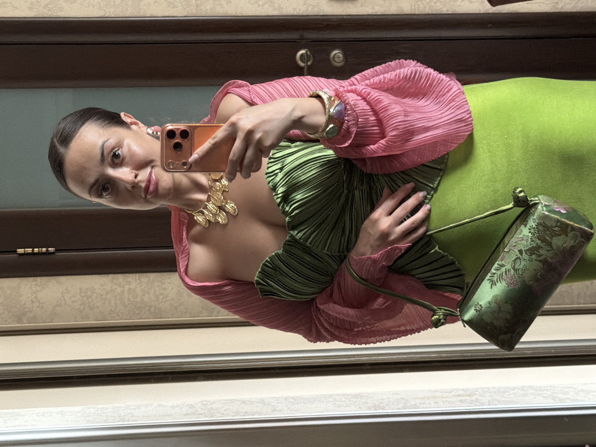 A woman with dark hair pulled back in a bun, wearing a green pleated strapless top and a long lime green skirt, is taking a mirror selfie. She is also wearing a pink pleated shawl, a gold necklace, a gold bracelet with colorful stones, and pink earrings. She is holding an orange smartphone in her right hand and a green patterned clutch bag in her left. The background appears to be an interior with wooden doors and trim, suggesting a home or hotel setting. The image does not provide enough visual information to determine the specific location within Krasnoyarsk, Russia.