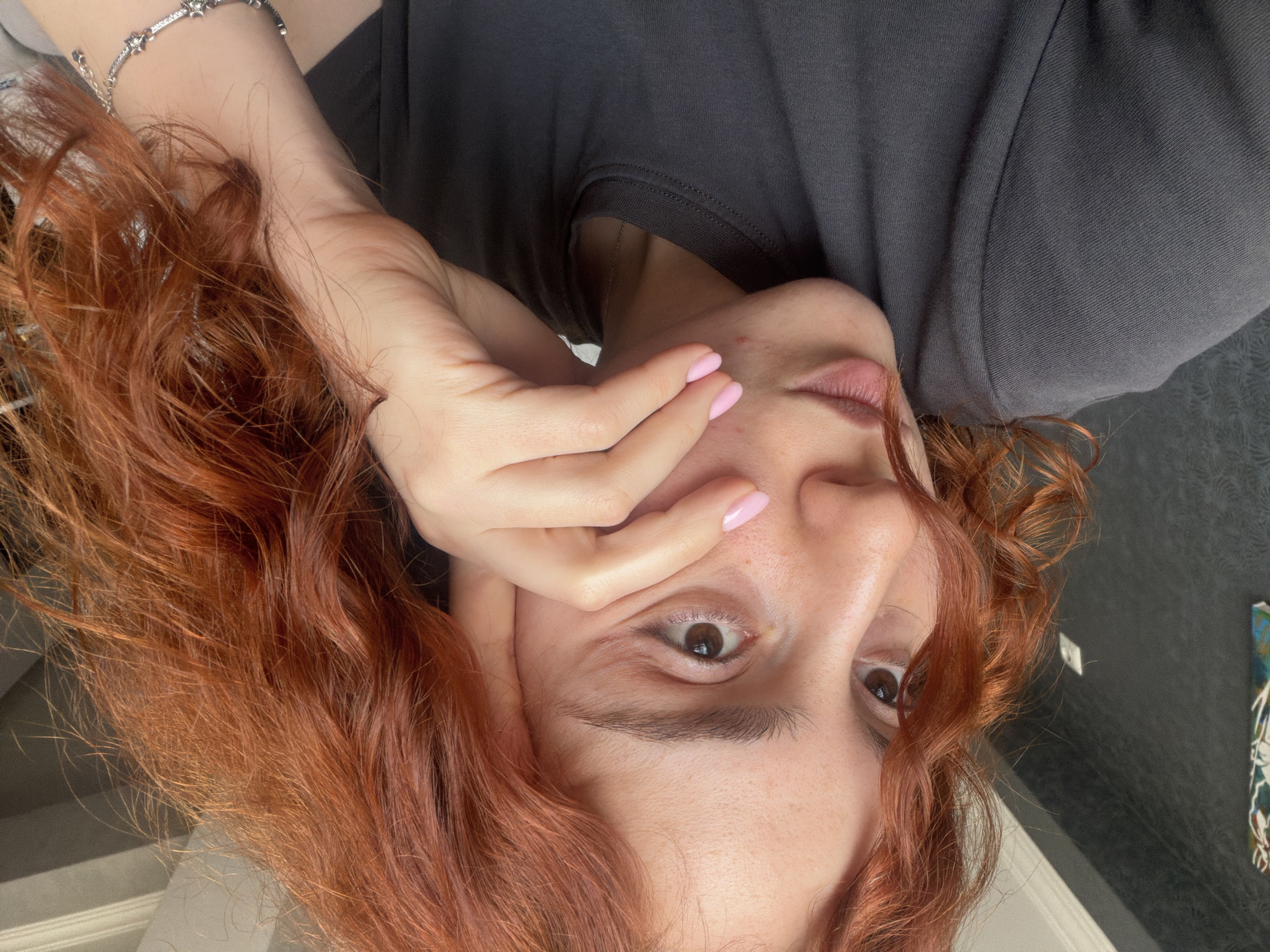 A woman with auburn, curly hair is positioned at an unusual angle, possibly reclining or tilting her head back. Her skin is fair, and she has visible eyebrows and brown eyes. Her left hand is partially obscuring her face, with nails painted a light pink color. She is wearing a dark gray t-shirt or sweater. The background is nondescript, with a dark-colored surface. The location of this image cannot be identified based on the image itself. It is unknown if this image was taken in Tbilisi, Georgia.