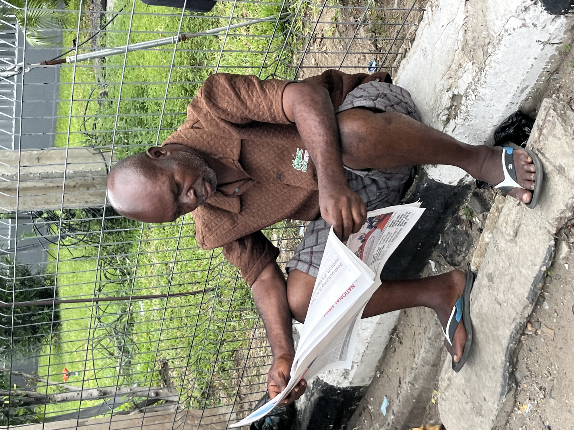 An adult male sits reading a newspaper outdoors in Itirin, Nigeria. The man is bald and dark-skinned, wearing a brown patterned shirt and plaid shorts. He sits on a concrete ledge next to a metal fence, reading a newspaper that reads "NATIONAL NEWS" at the top. The environment appears to be an urban area with foliage visible behind the fence.