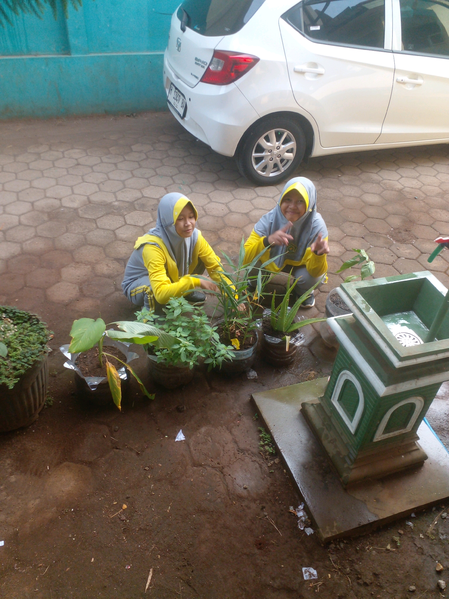 Two young Indonesian girls, wearing grey and yellow hijabs and matching athletic wear, are crouching in a yard. The girl on the left is tending to a potted plant, while the girl on the right is looking at the camera and gesturing with her hands. In the background, a white Honda Brio car is parked. The ground is paved with hexagonal stones and dirt. A green and white decorative structure with a shallow basin is visible in the foreground. The overall scene appears to be an outdoor area, possibly a schoolyard or residential courtyard, in Banyuputih, Indonesia, given the context of the car's license plate.