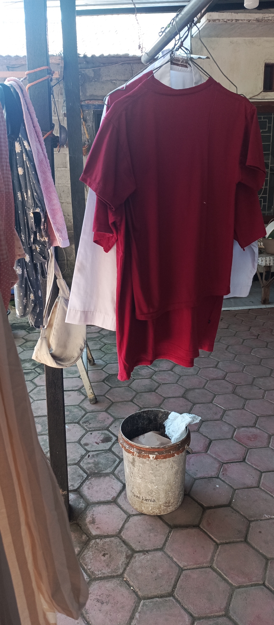 The image depicts an outdoor scene likely in the city of Arjasa, Indonesia, with laundry hanging on a clothesline. In the foreground, a cylindrical, weathered bucket with peeling paint contains some material and a crumpled white piece of paper. The bucket has the words "Kiwi Kimia" visible on its side. The ground is paved with hexagonal tiles in shades of red and gray. To the left of the bucket, a clothesline extends upwards, holding various items of clothing. Prominently featured is a bright red t-shirt, partially obscuring a white garment underneath. To the left of these, other clothes are visible, including a patterned dress with a pink towel draped over it. A cream-colored bag hangs from a support structure on the left side of the image. In the background, hints of a building with windows and a tiled roof are present, suggesting a residential or utilitarian outdoor area. The lighting suggests it is daytime, possibly late afternoon, given the shadows.