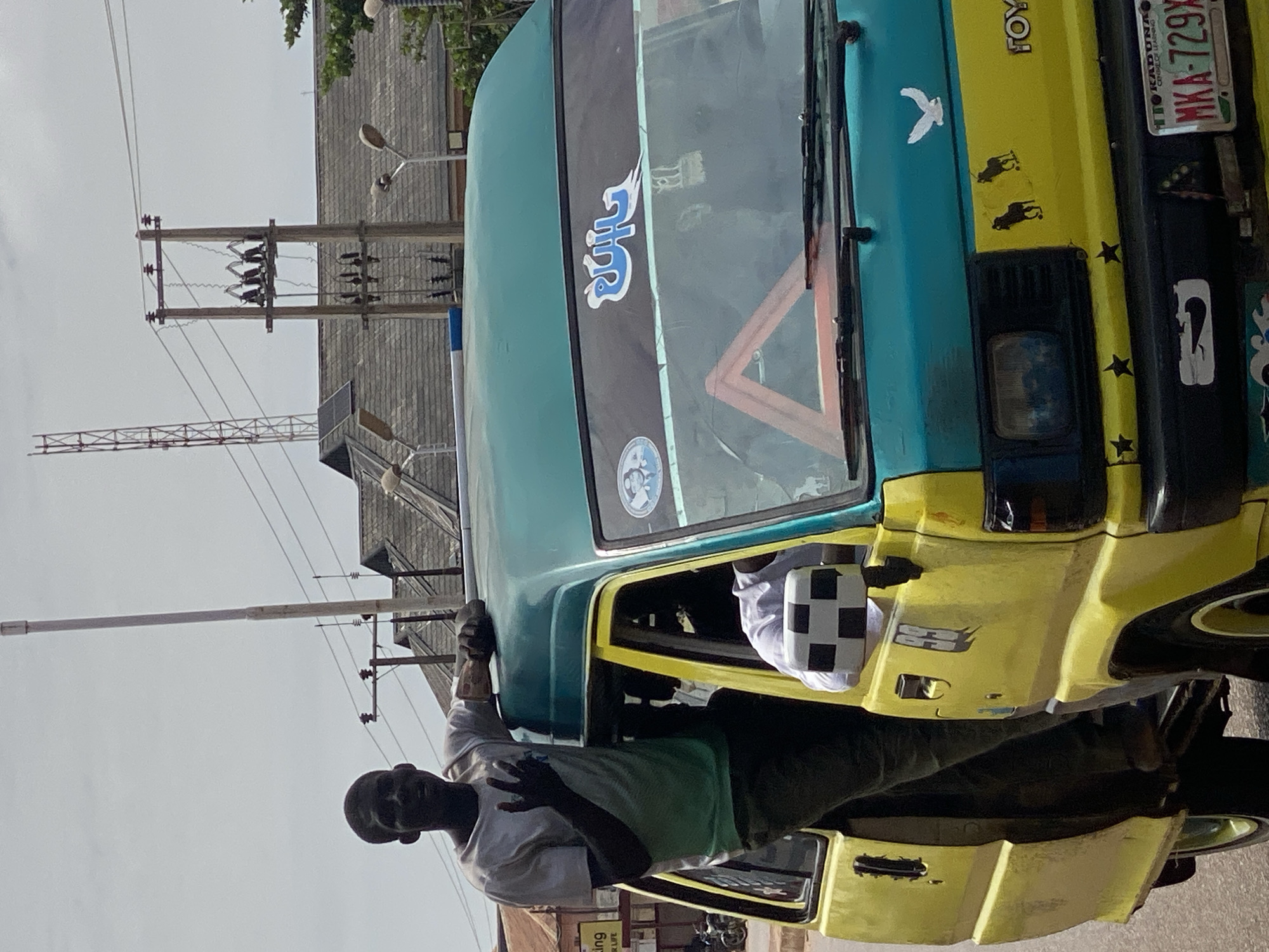 A Black man stands in the doorway of a brightly colored yellow and teal van, his left arm resting on the roof and his right hand raised in a gesture. He wears a light-colored t-shirt and dark pants. The van has several decals on its windshield, including a red triangular warning sign and a blue Arabic inscription. A license plate with the text "MKA-729X KADUNA" is visible on the front bumper. The background shows buildings with corrugated roofs, power lines, and a tall tower. The scene appears to be a street in Sabon Tasha, Nigeria, under an overcast sky.