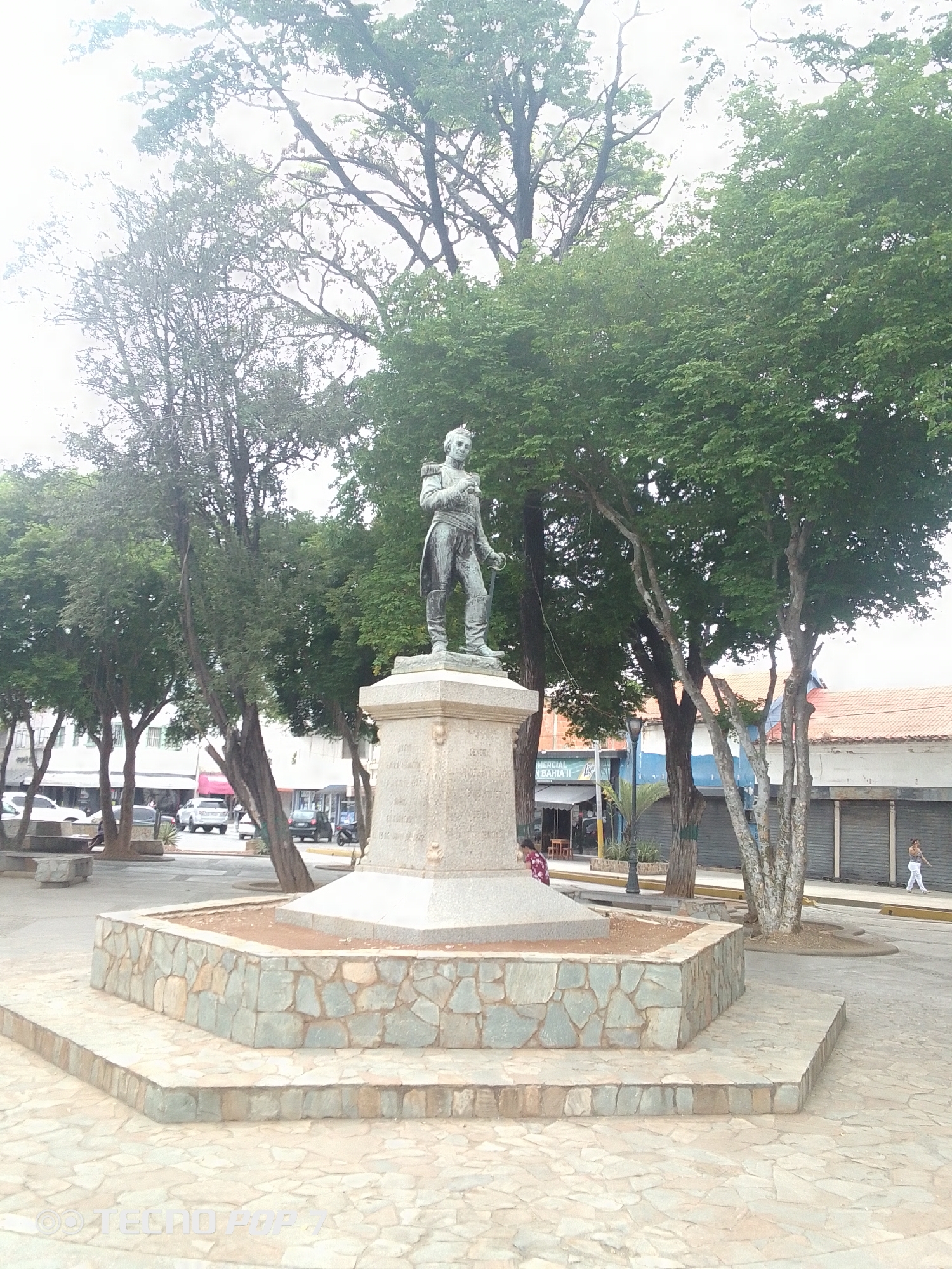 This image captures an outdoor scene in a public square or plaza, centered around a statue of a historical figure. The statue, made of a dark metal like bronze, depicts a man in military-style attire, complete with a jacket, epaulets, and a sash. He appears to be holding a scroll or document in his left hand and has his right hand on his chest. The statue stands on a multi-tiered stone pedestal, featuring a hexagonal base built from light-colored, irregularly cut stone blocks. Inscriptions, including the partially visible word "GENERAL," are carved into the main block of the pedestal.
The square is characterized by numerous green-leafed trees that create a lush, shaded environment. Their thick branches are visible between the clusters of foliage. The ground is paved with irregular, light-colored stone tiles. In the background, across a street with parked cars, stand several buildings, some of which appear to be storefronts with awnings and metal roll-down gates.
Two individuals are visible: one person in a patterned dress is partially obscured by trees near the statue's base, and another person in white pants and a light top walks along the sidewalk in the far right background.
The scene takes place during the daytime, indicated by the bright, diffused lighting, suggesting an overcast sky. The overall atmosphere is calm and ordinary, depicting daily life in a public space. A "OO TECNO POP 7" watermark is present in the bottom-left corner. The setting is consistent with a town square in Altagracia, Venezuela.