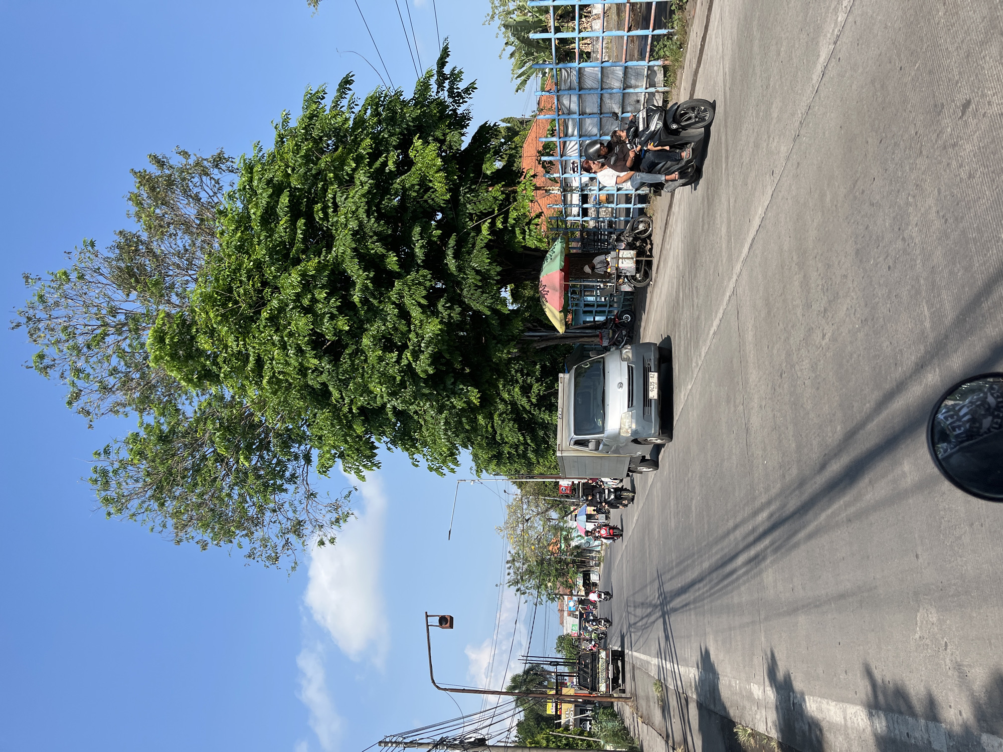 The image depicts a street scene in Tulangan, Indonesia, under a clear blue sky with scattered clouds. The perspective is from a moving vehicle, likely a motorcycle given the reflection in the lower frame. The road is paved with asphalt and marked with a white line on the left side. Several vehicles are visible: a silver van in the foreground to the right, a black pickup truck further down the road on the left, and numerous motorcycles carrying one or two passengers.
On the right side of the road, a blue fence borders a property with trees and some buildings behind it. A colorful umbrella shades a vendor stall where a person stands next to a motorcycle. A man and a child are riding a black motorcycle towards the viewer on the right side of the road.
The left side of the road features a sidewalk with some vegetation, utility poles with numerous wires, and some small businesses. A traffic light hangs over the road. Tall trees with dense green foliage dominate the upper and right portions of the image, casting shadows on the road. The overall impression is a moderately busy street in a tropical climate.