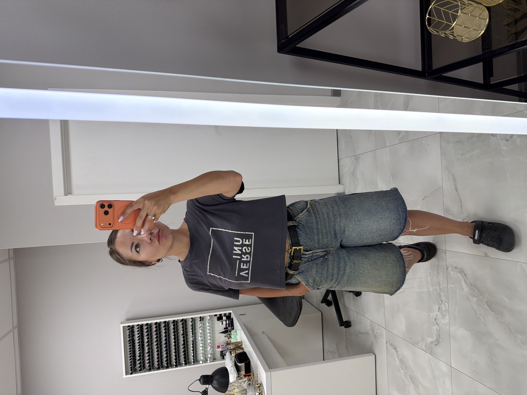 A woman stands in a well-lit room, taking a selfie in a full-length mirror. The room appears to be a nail salon, indicated by a white desk with various nail polish bottles and tools arranged on it, along with a rack displaying rows of nail polish. She is wearing a dark gray, cropped t-shirt with a white graphic print and denim shorts. She has a black belt with a gold buckle. Her hair is pulled back, and she is wearing hoop earrings and rings. She is holding an orange smartphone to take the picture. The floor is tiled with a marble pattern. To the right of the mirror, a tall, thin, illuminated column is visible, as is a black metal shelf unit with a gold decorative basket on it. The woman is looking directly at her reflection in the mirror.