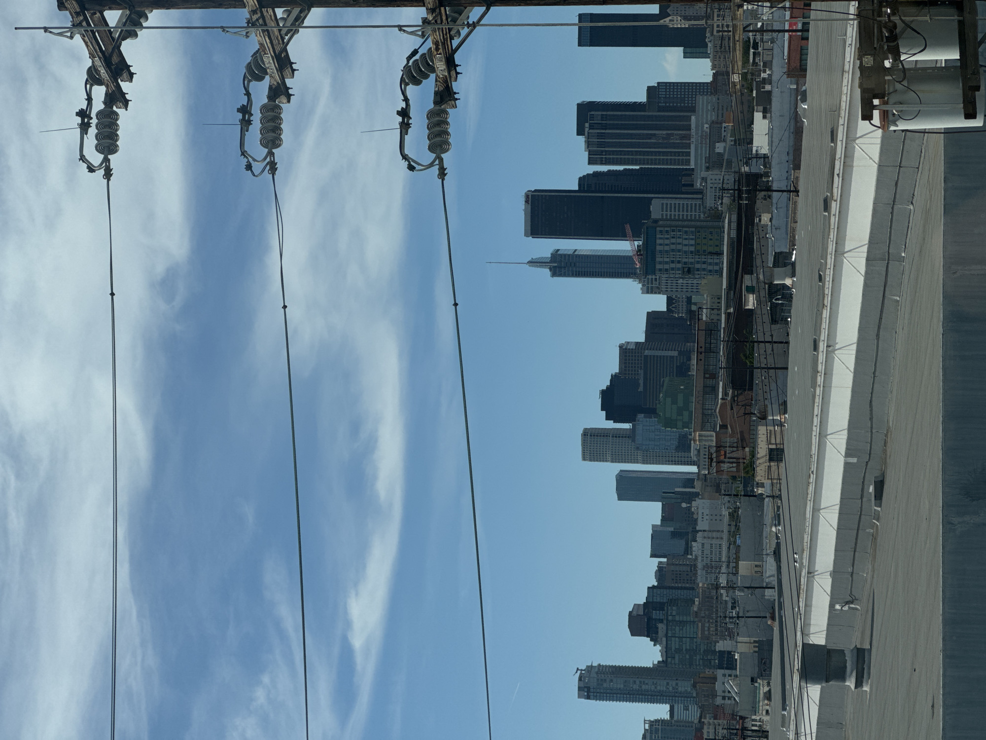 The image shows a view of the Los Angeles skyline, likely from a rooftop. Power lines and utility poles are visible in the foreground. The background features numerous skyscrapers of varying heights, suggesting a downtown or urban area. The sky is blue with scattered clouds. The angle of the photo suggests that it was taken with the camera turned sideways.