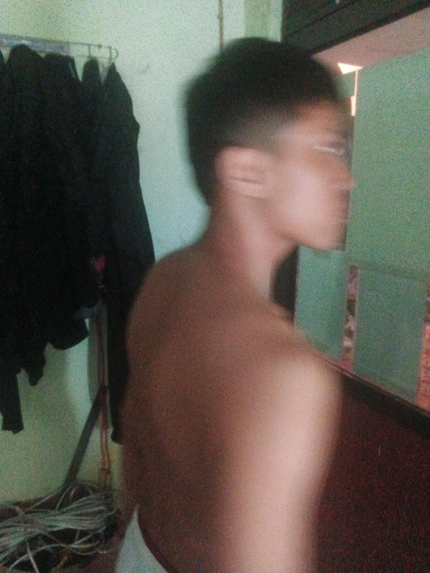 The image shows a blurred indoor scene. In the foreground, a person is seen from the side, shirtless. They have short dark hair and appear to be wearing glasses. Their back and shoulder are visible. In the background, there are dark garments hanging on a rack on the left. The walls are a light green color. On the right, there is a dark surface, possibly furniture, and a green wall with some patterned elements. The overall lighting is dim. The location is specified as Keji, Indonesia, but there are no visible elements within the image that definitively confirm this location.