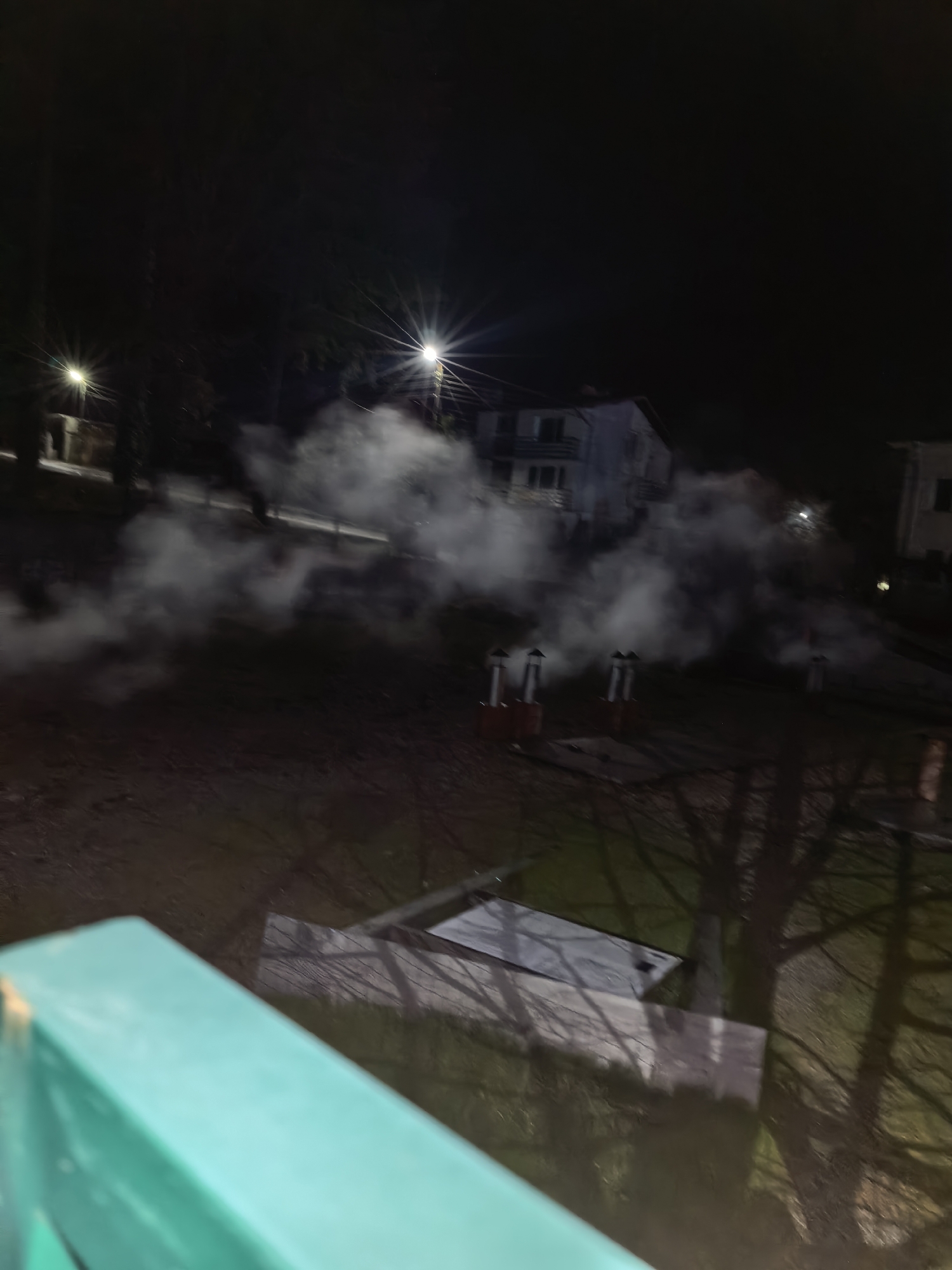 The image, taken at night, depicts a scene in an urban area of Unknown, Unknown. In the background, there is a building that appears to be residential, possibly a multi-story house. Streetlights illuminate the scene, and there is visible smoke or steam in the air, obscuring the view. Several red objects with chimney-like structures are visible, seemingly emitting the smoke. In the foreground, there is a glimpse of what appears to be a rooftop or balcony, with bare tree branches visible. The image is somewhat blurry, but the overall impression is of a night scene in a residential area, with visible signs of smoke or steam emission.