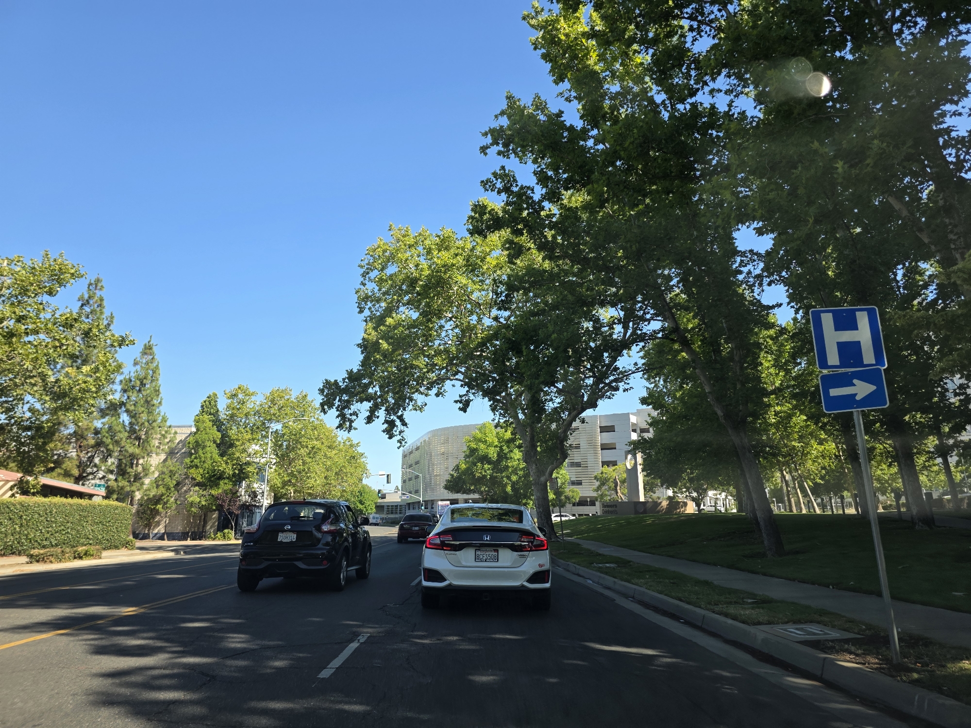 The image depicts a scene from a vehicle on a sunny day, likely in Sacramento, California. The sky is a clear, bright blue, suggesting good weather. The setting is a suburban or commercial area with mature trees lining the street.
In the foreground, a white Honda Civic is driving in the right lane, with its rear visible. Ahead of it, a black Nissan Juke is also driving in the same lane. To the left, another vehicle, partially obscured, is visible in the left lane. The road is paved with asphalt and has clear lane markings, including yellow lines separating opposing traffic and white lines marking the lanes.
On the right side of the road, a sidewalk runs alongside a grassy area with several large trees. A blue sign with a white "H" symbol and a white arrow pointing to the right is mounted on a pole. Further down the street, a modern building with a curved, white facade is visible, appearing to be a commercial or institutional structure. A sign at the base of a monument reads "Sutter's Gardens."
The time of day appears to be late morning or early afternoon, given the bright sunlight and the shadows cast by the trees. The overall mood is peaceful and ordinary, depicting a typical day of driving in a well-maintained urban environment. No people are clearly discernible in the image, as the focus is on the road and surrounding environment.