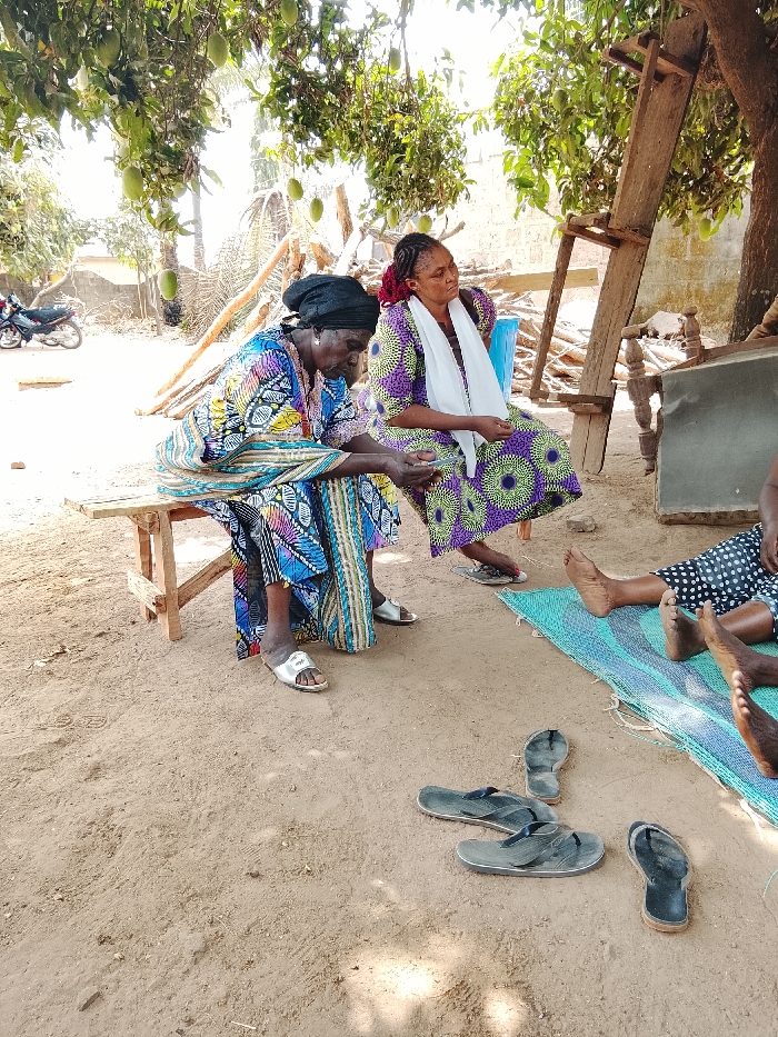 The scene is an outdoor gathering in Akwanga, Nigeria, under the shade of trees bearing unripe mangoes. The ground is dry, light brown soil.
Three individuals are present. To the left, an elderly woman with dark skin and a black headwrap sits on a light wooden bench. She wears a long, colorful patterned dress in shades of blue, green, and yellow, and silver slide sandals. Her hands are clasped in front of her, and her gaze is directed downward. To her right, a younger woman sits on a light blue plastic chair. She wears a purple and green patterned dress, a white scarf around her neck, and her hair is styled in braids. She looks towards the right. To the far right, the bare lower legs and feet of at least one other person are visible, resting on a blue and green woven mat laid on the ground. This individual wears a dark garment with white polka dots.
On the ground in front of the mat, three pairs of dark-colored flip-flops are scattered. In the background, to the left, a red and black motorcycle is parked near a light-colored wall. Further back and to the right, a tall wooden structure, possibly a ladder or scaffold, leans against a tree. Overhead, numerous green, unripe mangoes hang from the leafy branches. The lighting suggests bright daylight.