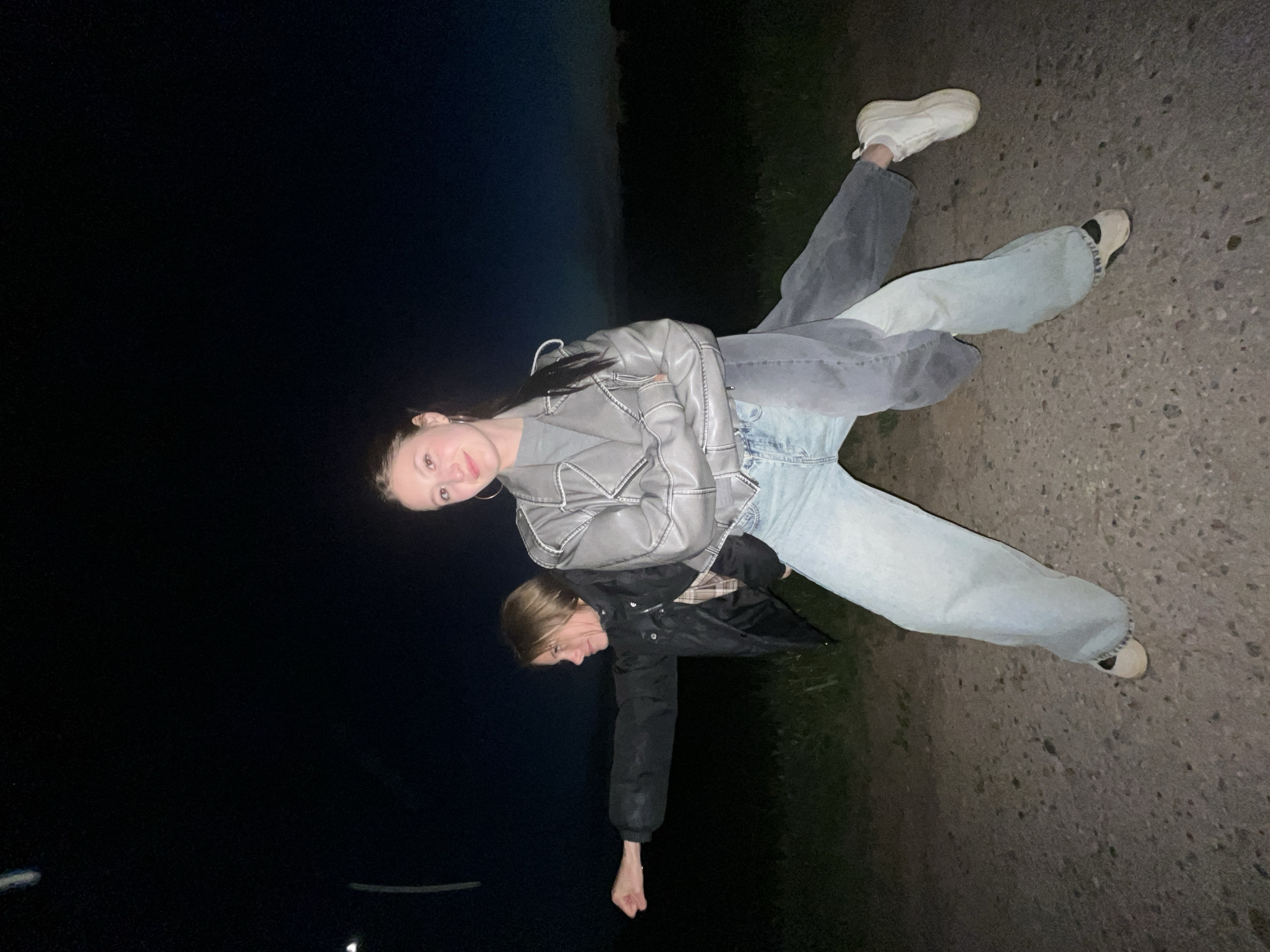 Two young women are visible in the image. The woman on top wears a grey leather jacket and light blue jeans. The other woman wears a dark jacket. They are positioned on what appears to be a road or paved area at night. The backdrop consists of a dark sky and an unlit area, suggesting a rural or suburban location. The location in the city of Фоки, Russia, cannot be determined from the image.