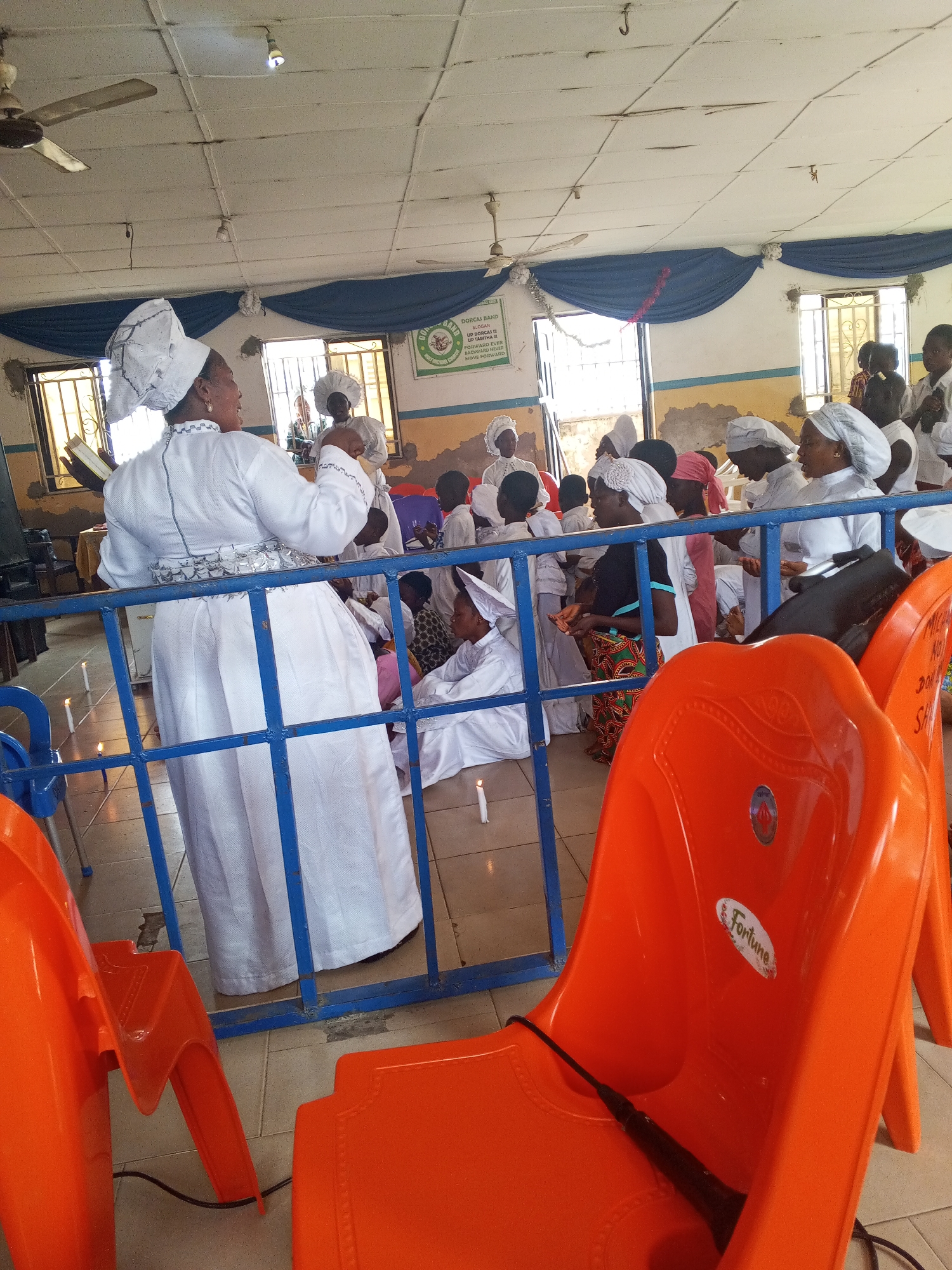 A group of people, mainly women and children, are gathered in a brightly lit indoor space, possibly a church or religious gathering place, in Lokoja, Nigeria. The focus is on a woman standing behind a blue metal railing, wearing a white garment and headwear, and holding a book, as if addressing the group. Many others in the assembly are also dressed in white attire with distinctive headwear.
Several orange plastic chairs, with the brand name "Fortune" visible on one, are in the foreground. The interior features windows, ceiling fans, and blue fabric draped as decoration. A sign with the words "DORCAS BAND" is visible on the wall. Lit candles are placed on the floor around the group, adding to the solemn atmosphere. The overall impression is of a religious ceremony or meeting taking place.