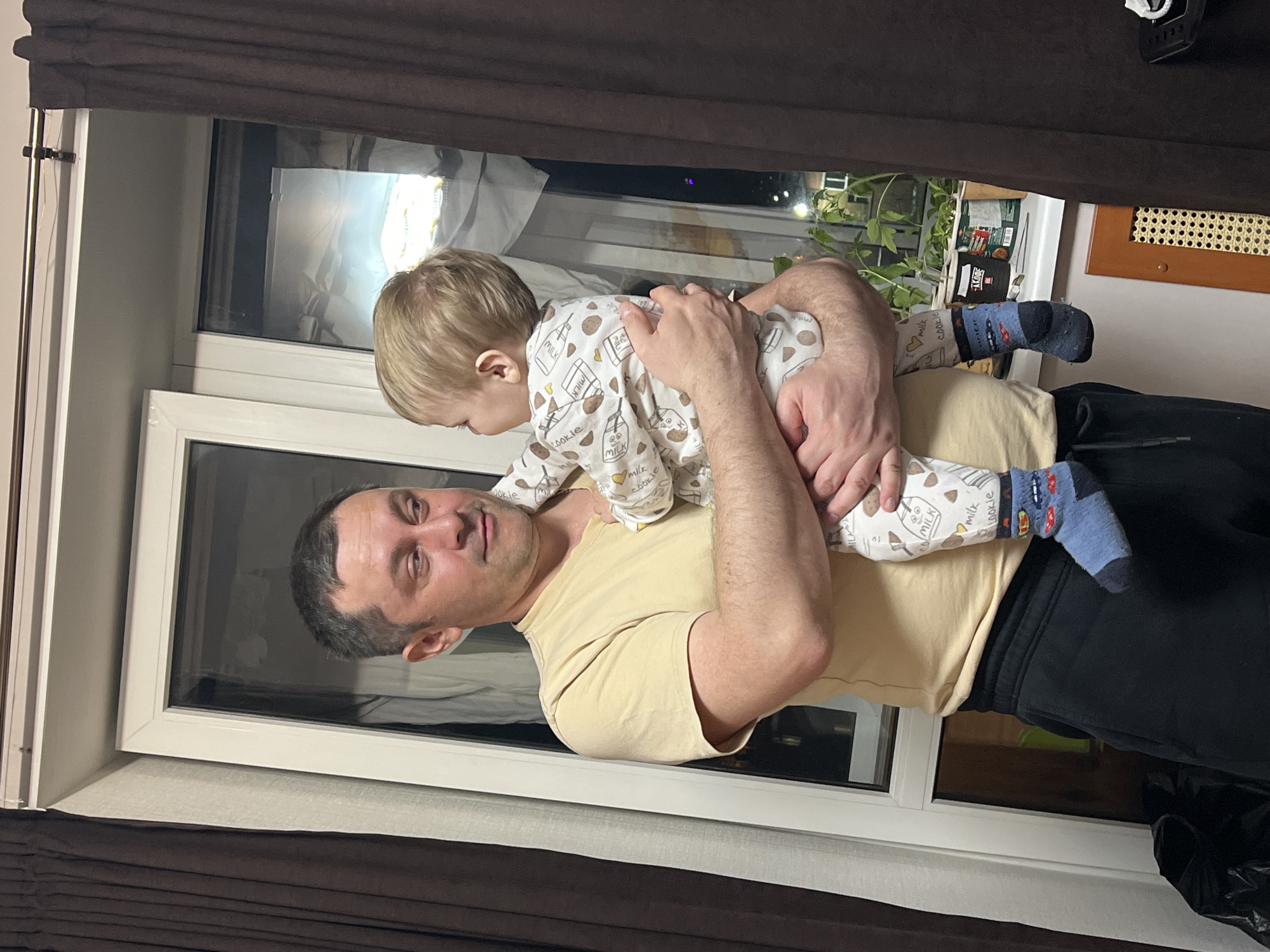 This image, captured in Perm, Russia, depicts a heartwarming indoor scene featuring an adult man holding a young child. The man, with short dark hair, wears a light yellow t-shirt and dark trousers, looking directly at the camera with a calm, content expression. He cradles the child, who has light blonde hair and is dressed in a white onesie adorned with patterns of milk cartons and cookies, alongside visible text like "MILK" and "COOKIE." The child is also wearing blue socks featuring car designs and is positioned across the man's torso, facing away from the camera.
The setting appears to be a home, with a prominent white-framed window in the background. The view through the window indicates it is nighttime, as distant artificial lights are visible against a dark sky. Dark brown curtains or blinds partially frame the window at the top and bottom. On a ledge or windowsill outside the window, a green houseplant can be seen, along with other obscure objects, one of which displays the text "K CORE." To the right of the window, a wooden grate, possibly for ventilation, is visible on the wall. A metallic rod is present on the left side, likely part of the window or curtain fixture. The overall scene conveys a tender, familial moment.