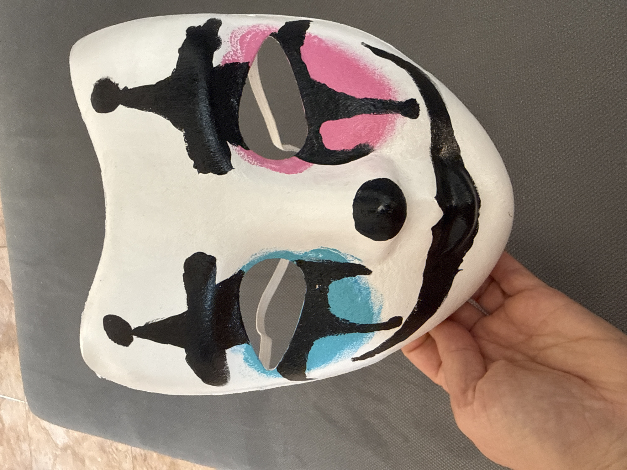 A hand holds a white face mask with black, pink, and blue markings. The mask has two eyeholes filled with gray material. The mask rests against a gray surface.