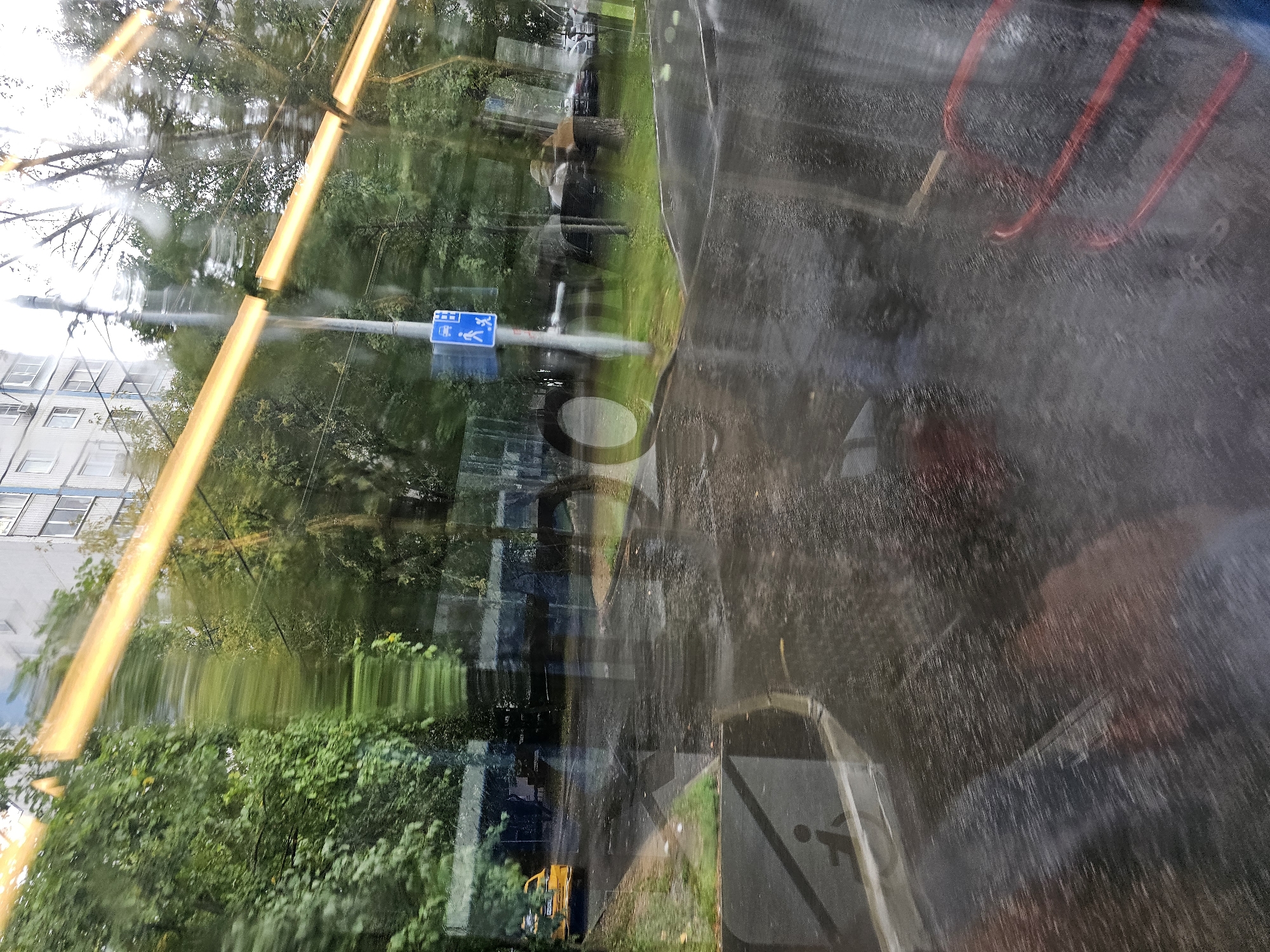 The image depicts a view through a window, likely from a vehicle, looking out onto a wet, overcast street scene in Khimki, Russia. The glass is streaked, and there are reflections on its surface.
In the foreground, the dark, wet pavement reflects the light from overhead. Large, blurry, circular shapes appear on the surface, possibly reflections of light fixtures or structural elements. To the left, a portion of a sidewalk or pedestrian area is visible, featuring a pedestrian crossing sign with a silhouetted figure.
Further back, a yellow vehicle, possibly a taxi, is parked or driving on the left side of the road. Trees with dense green foliage line the street. A modern, multi-story building is visible in the background, with multiple windows. A traffic light pole with a blue pedestrian and bus crossing sign is positioned near the center of the frame. The overall impression is that of a city street after rain. The reflections on the window are significant, obscuring some details of the external scene. Red, curved objects, possibly seating or railing, are visible in the lower right quadrant of the image, also distorted by reflections.