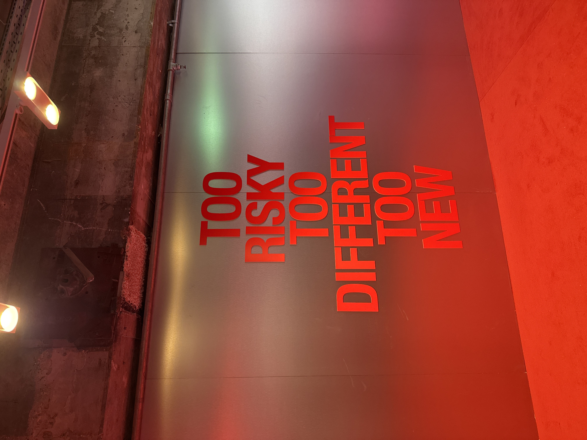 The image presents a wall with red text on a silver background stating "TOO RISKY TOO DIFFERENT TOO NEW." The wall is illuminated by lights on the left side, casting a glow. To the left of the wall, there is a textured, gray, concrete-like structure with some exposed elements. Adjacent to the right side of the wall is a reddish-orange surface. Without further context, it is impossible to determine the exact location in London, United Kingdom.