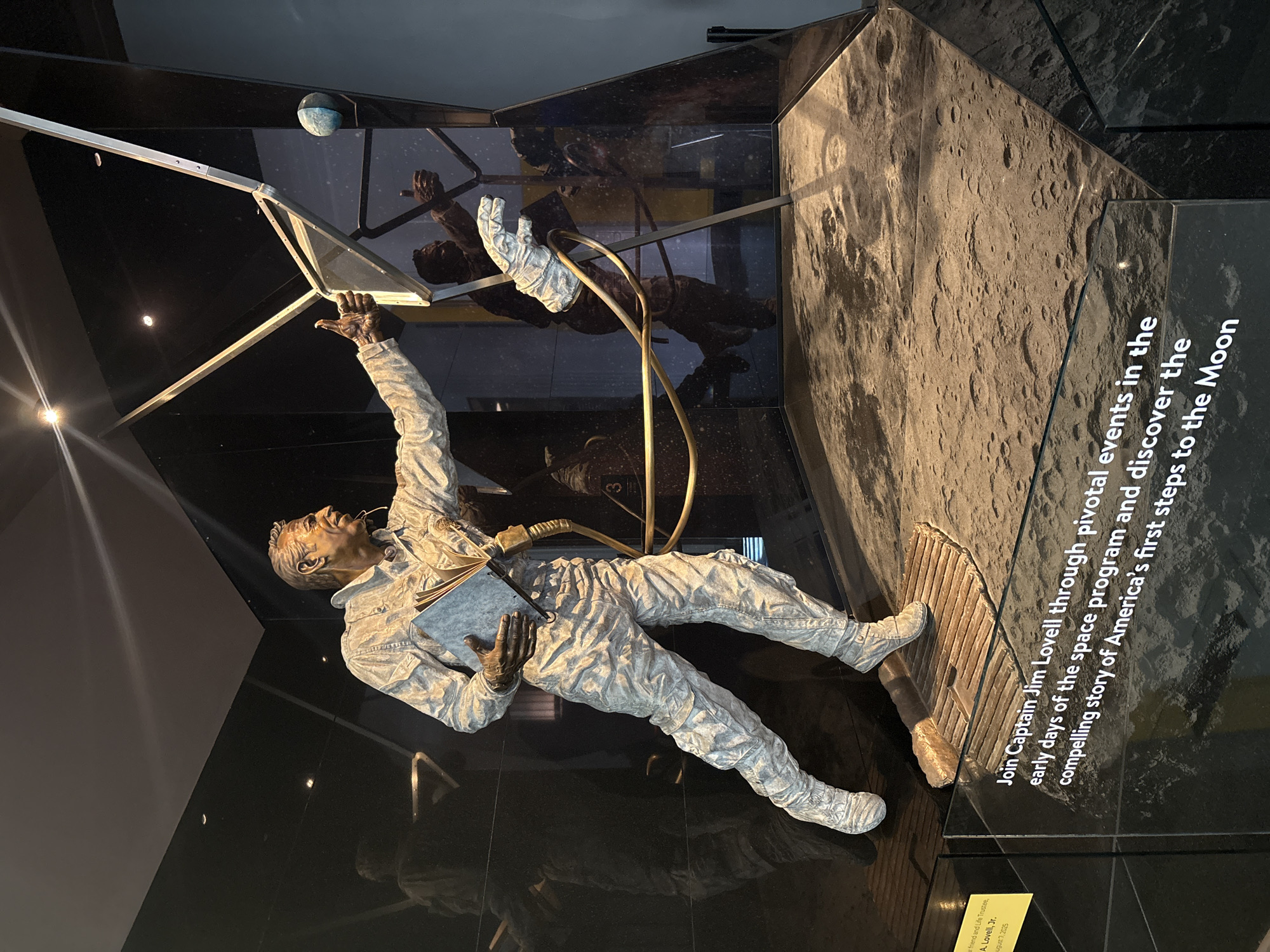 A photograph shows a display, likely inside a museum, featuring a statue of Captain Jim Lovell. He is dressed in a space suit, holding a book and a pen, reaching towards a structure above him that resembles a lunar lander. The statue is positioned inside a display case with black walls and a glass front. A section of the display case wall has a textured surface resembling the moon's surface. Text on the display reads: "Join Captain Jim Lovell through pivotal events in the early days of the space program and discover the compelling story of America's first steps to the Moon". Also, a small yellow card to the bottom of the photograph reads: "A friend and life trustee, A. Lovell, Jr., August 7, 2025".
While the exact location is not explicitly stated, the presence of a tribute to Captain Jim Lovell and the focus on the space program suggests it is likely a museum or exhibition in Chicago, United States of America.