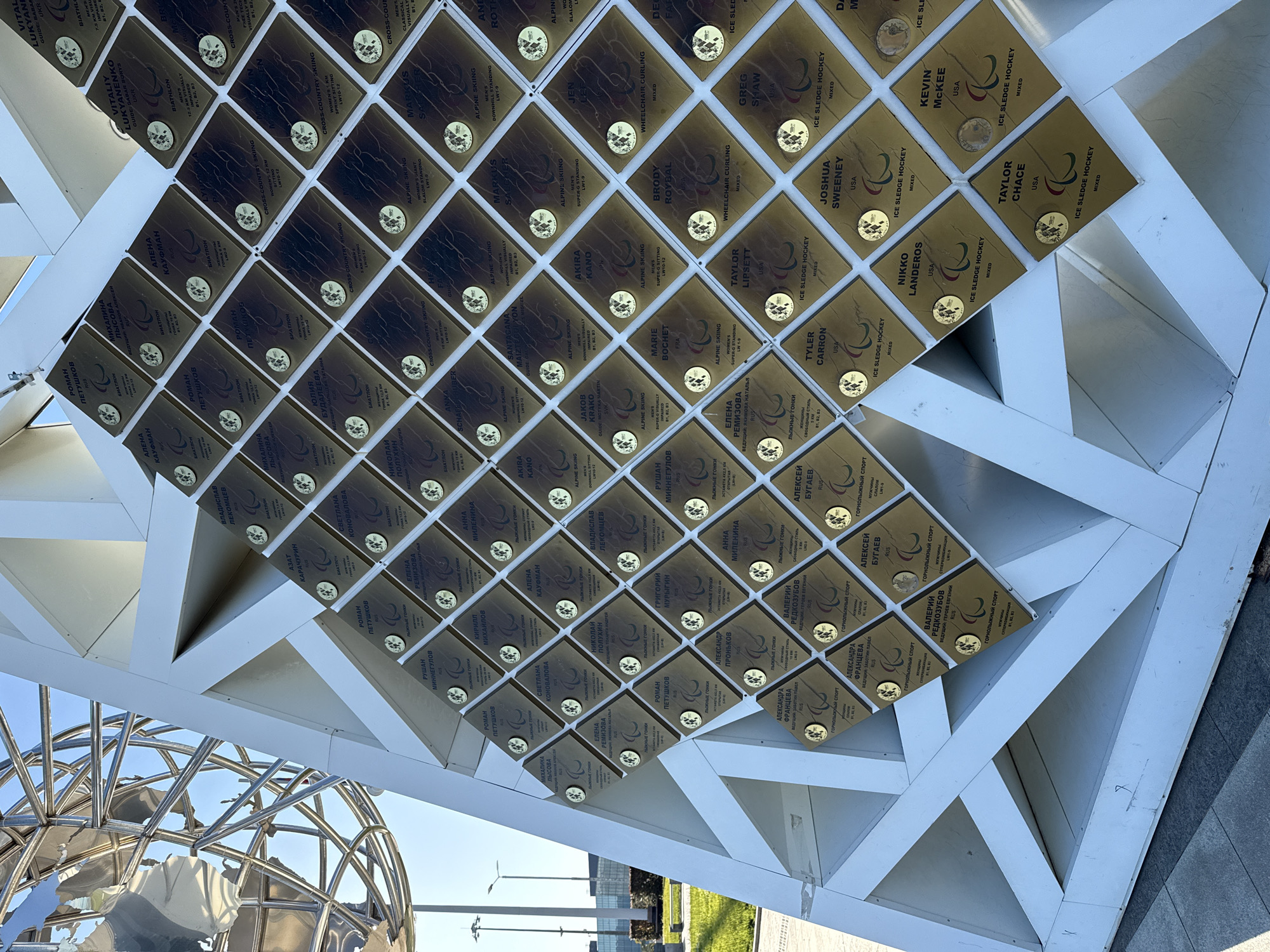 This image shows a structure adorned with plaques bearing the names of Paralympians. The plaques are mounted on a grid-like framework composed of white beams. Each plaque features a name, sport, and country, along with a circular emblem. The sports include biathlon, cross-country skiing, alpine skiing, and ice sledge hockey. Notable countries represented are Russia, USA, Ukraine, France, and Japan.
The location is identified as the city of Sirius, Russia. The structure appears to be an outdoor memorial or installation, with partial views of a spherical metal sculpture, pavement, and greenery. The lighting suggests daytime under a clear sky.