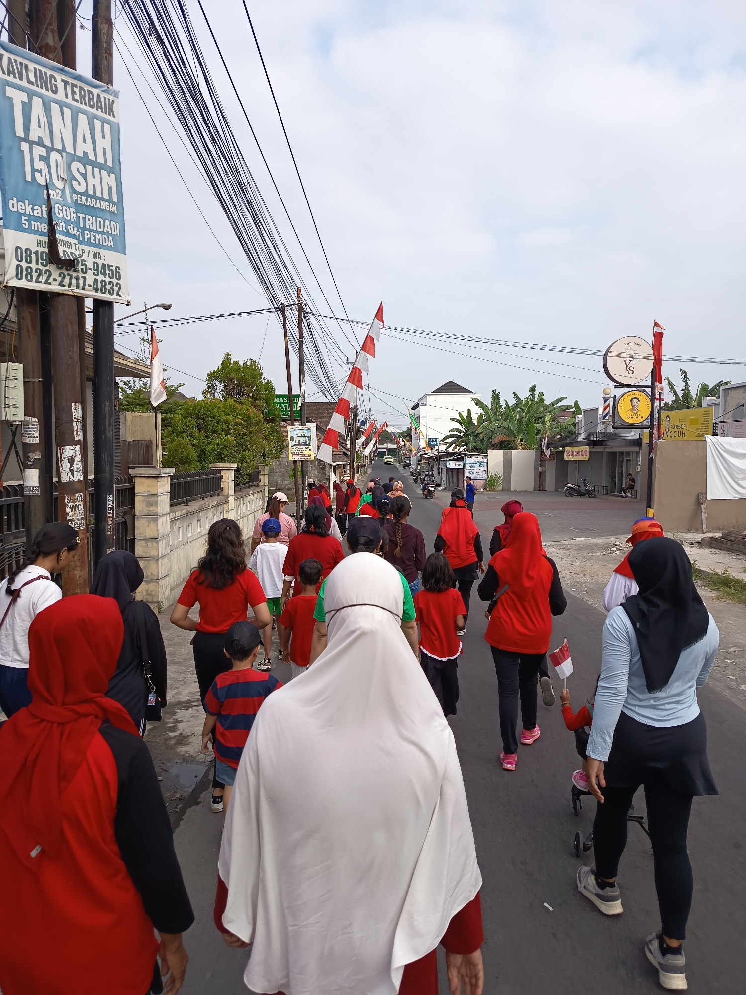 In Ngaglik, Indonesia, a group of people, predominantly wearing red and white attire, walk along a paved road under an overcast sky. Indonesian flags are visible, some attached to poles and others held by individuals. The group includes adults and children, with some children walking alongside or being pushed in strollers. The surrounding environment features buildings, streetlights, and power lines, suggesting a semi-urban setting. Signs in Indonesian are present on buildings and poles, indicating commercial establishments and advertisements. The people appear to be participating in an outdoor event or procession.