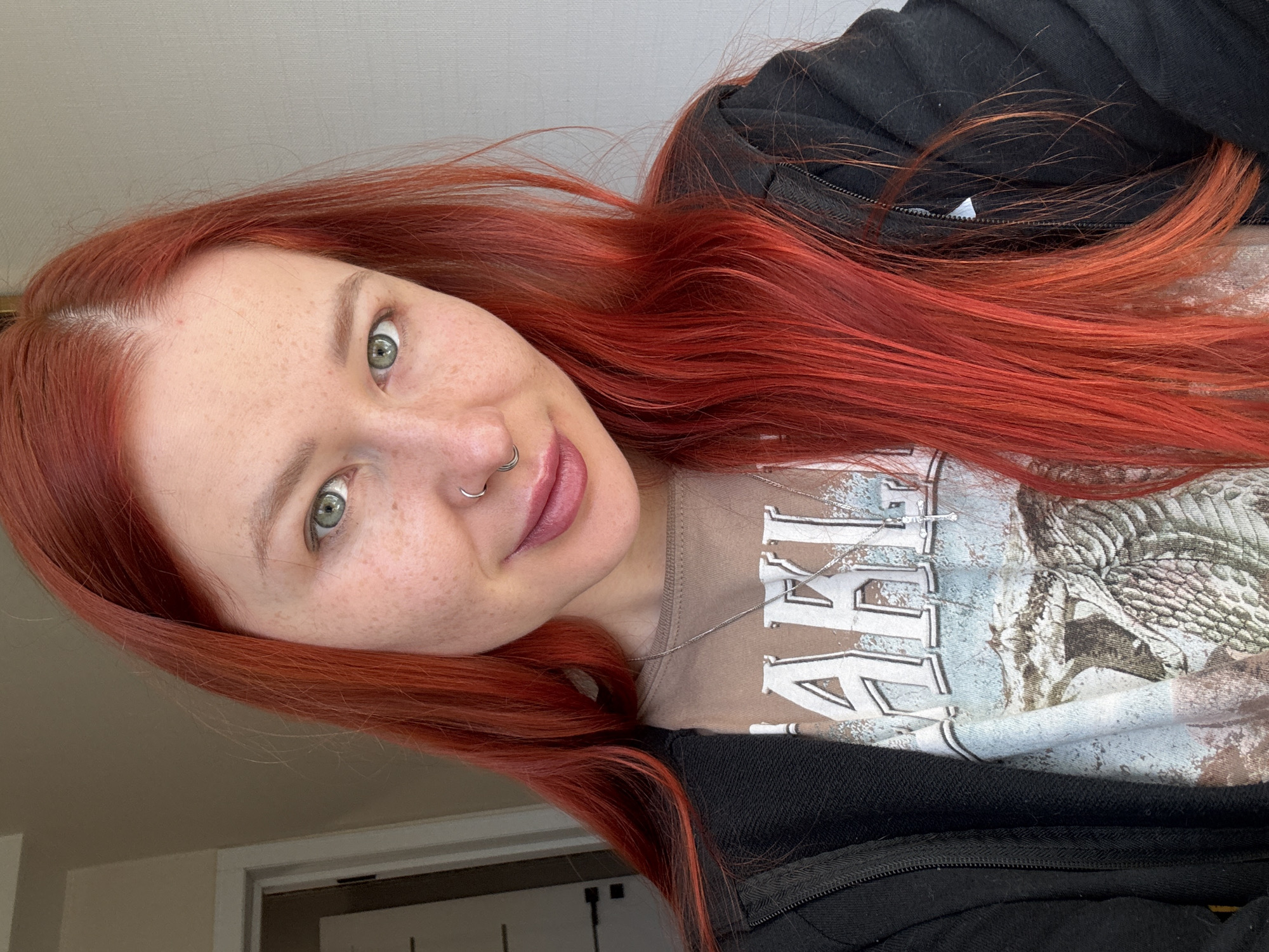 A young woman with vibrant, long red hair and green eyes poses for a close-up photo, tilting her head. Her skin is pale and adorned with numerous freckles across her nose and cheeks. She has a slight, soft smile on her lips, which are coated with a light pink lipstick. A silver hoop septum piercing is visible on her nose.
She is wearing a dark, possibly black, zipped hoodie over a light brown or tan t-shirt. A thin silver chain necklace is visible around her neck, resting on the t-shirt. The t-shirt features a large graphic print that includes what appears to be a bird, possibly an eagle or hawk, with detailed feathering. Parts of stylized text are also visible on the shirt, notably "RE" in block letters, suggesting a band name or similar design.
The background is simple and uncluttered, consisting of a plain, light-colored wall or ceiling in the upper part of the frame, and a glimpse of a door frame on the lower left. The lighting is bright and even, indicating daytime. The overall setting appears to be indoors, possibly a casual home environment. The image is taken in Rostov-on-Don, Russia.