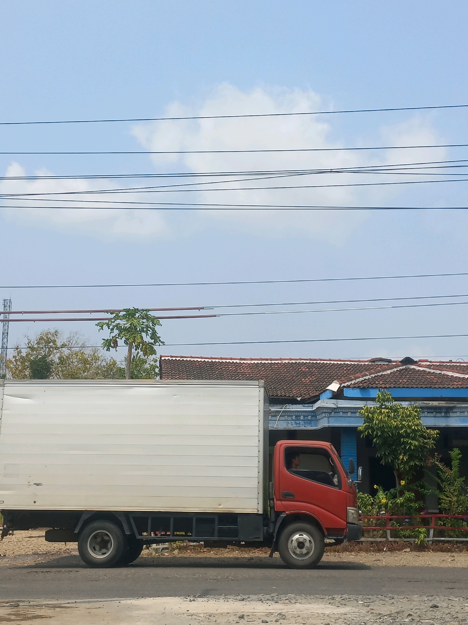 An orange and white delivery truck is parked on the side of a road in Baturetno, Indonesia. The truck has a white cargo box and an orange cab, with a person sitting in the driver's seat. Behind the truck, a blue house with a tiled roof is visible, surrounded by trees and foliage. Several power lines are strung across the sky, which is mostly clear with a few scattered clouds. The road in front of the truck is paved with asphalt and gravel.