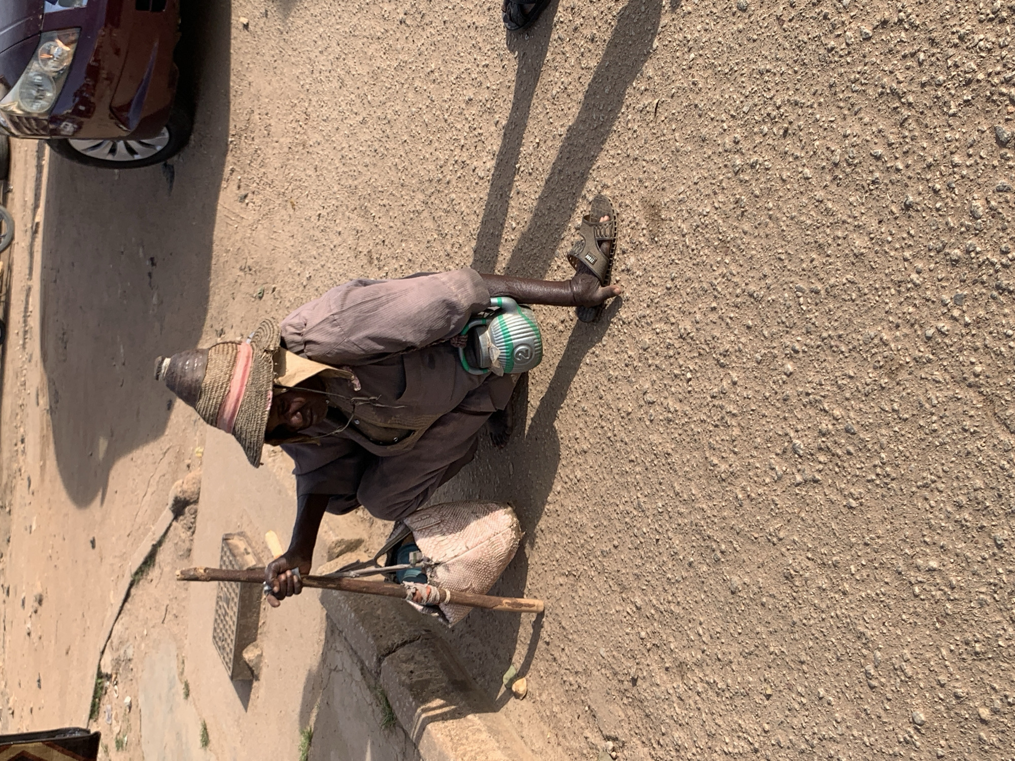 A person wearing a straw hat and a long-sleeved garment is crouching on a dirt surface in Jos, Nigeria. The person holds a wooden stick in their right hand, and a light-colored bag with a textured pattern rests beside them. A small, green and white jug with a handle is positioned in front of their right leg. The person is also wearing sandals on their feet. The ground is covered in loose dirt and small stones, with a concrete curb visible to the left. A shadow of a person is cast on the ground. A portion of a dark-colored vehicle is visible in the upper right corner of the image. The scene appears to be outdoors, possibly on a street or roadside.