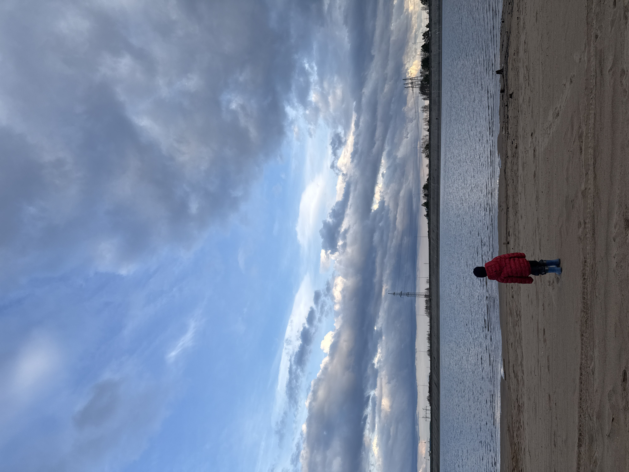 The image captures a serene outdoor scene by a body of water, located in Noviy, Russia. The foreground features a sandy bank with visible footprints. A child, dressed in a red puffer jacket, dark hat, and blue boots, stands with their back to the camera on the sandy shore, looking towards the water. The calm surface of the water occupies the midground, reflecting the subdued light from the sky.
The sky is a prominent feature, filled with dramatic, elongated grey clouds that stretch across the expanse, interspersed with patches of bright blue sky where sunlight breaks through. In the far background, across the water, a thin line of trees is visible, along with several tall utility poles, suggesting a distant industrial or infrastructure presence.
The lighting indicates either a late afternoon, early evening, or a heavily overcast day, with the sun low on the horizon, creating a soft and contemplative atmosphere. There is no visible text in the media file.