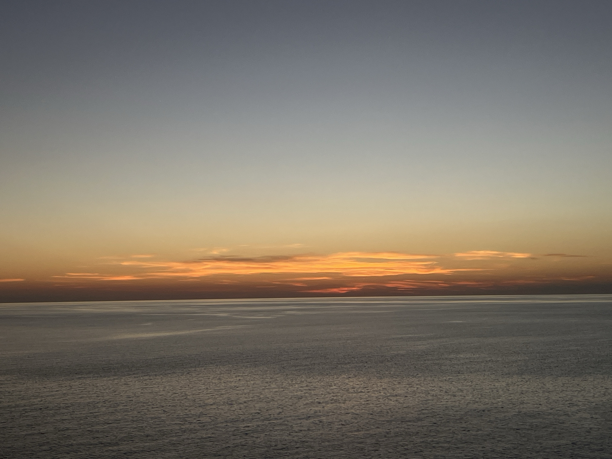 The image depicts a seascape at dusk or dawn. The sky transitions from a light grey-blue at the top to a band of orange and yellow hues near the horizon, where a few clouds are visible. The sea occupies the majority of the frame, displaying a muted grey tone with subtle ripples on the surface. The horizon line is visible in the middle of the image. Location: Unknown, Unknown.