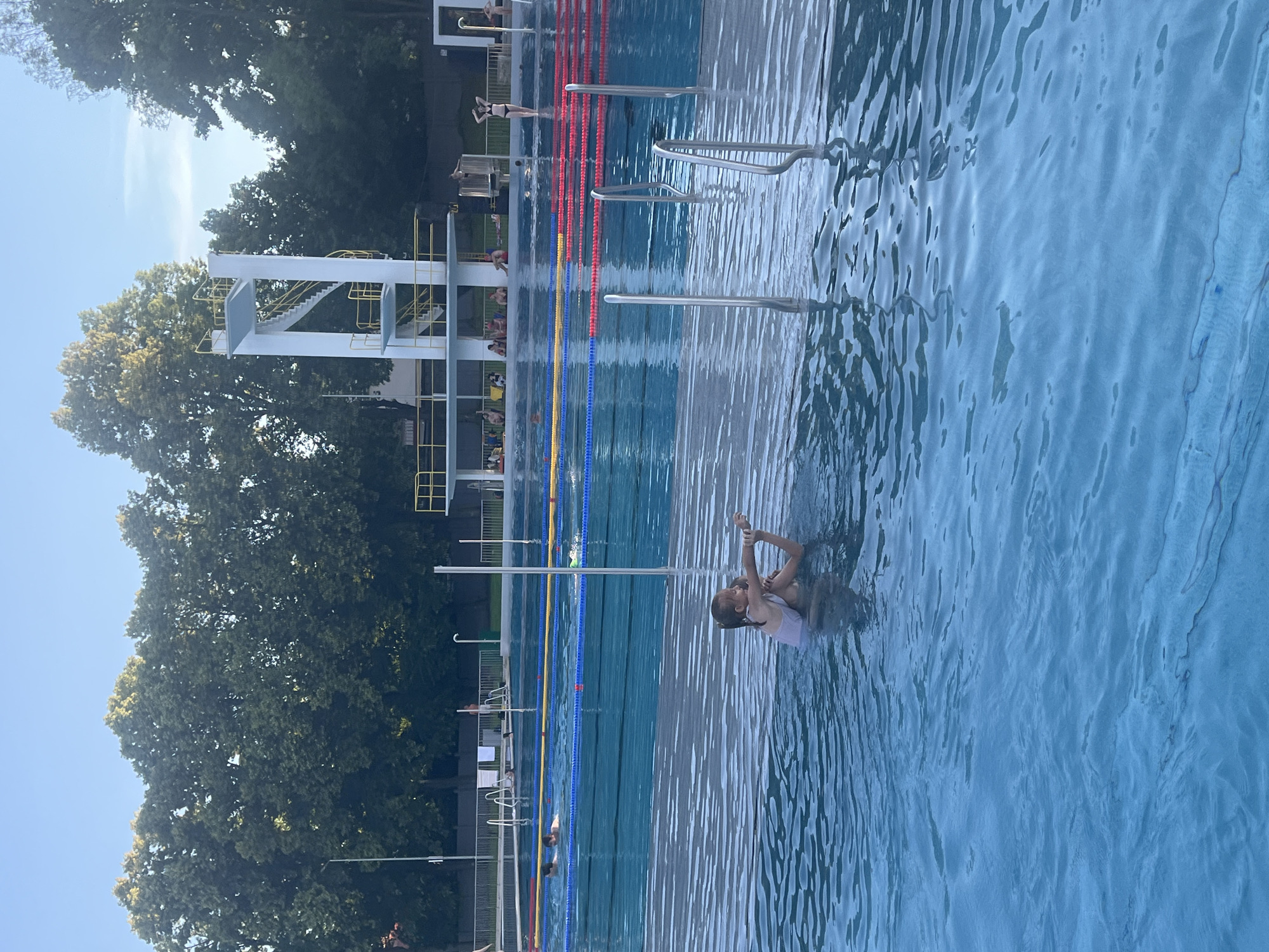 In an outdoor public swimming pool, a young girl with blonde hair, wearing a light-colored swimsuit, is being held by an adult in the shallow end of the water. The adult is supporting the girl, who appears to be performing a pose or movement in the water. Several swim lanes are visible in the background, marked by buoys and lane lines in red, yellow, and blue. Further back, a diving platform with a white structure and yellow railings stands against a backdrop of lush green trees under a clear blue sky. Other people are seen scattered around the pool area, some swimming in the lanes and others relaxing or observing from the poolside. The water surface is slightly rippled, reflecting the sky and surrounding structures.