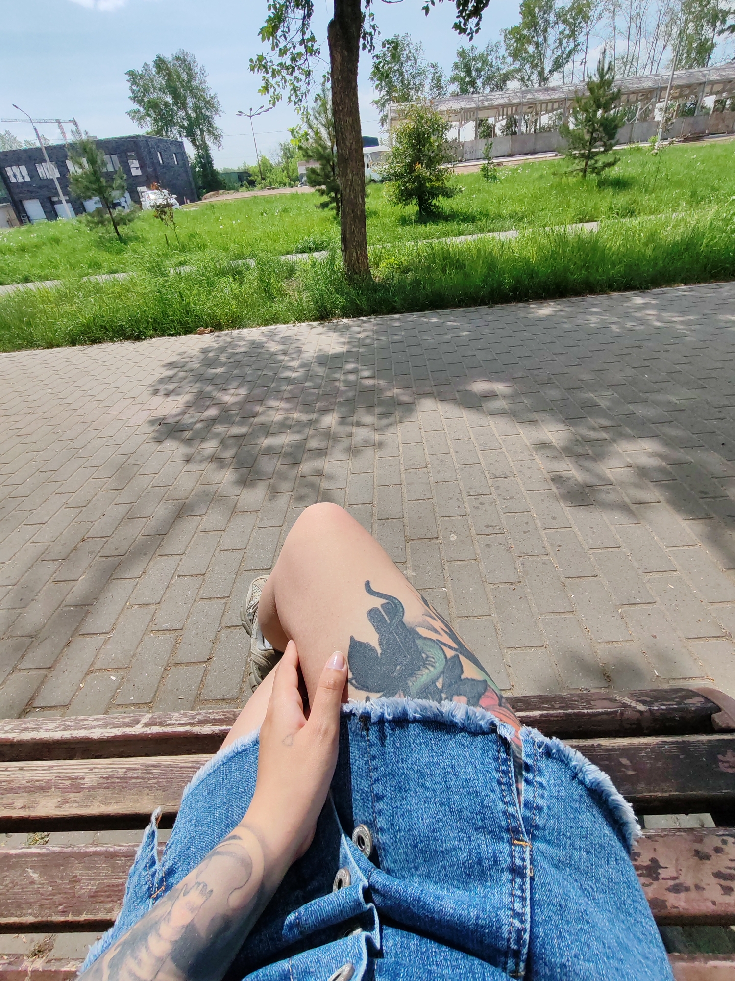 A low-angle, eye-level photograph depicts a person sitting on a wooden bench in a park or outdoor area in Chaikovsky, Russia. The image is shot from the person's perspective, showing their lap, legs, and parts of their arms. The individual is wearing a blue denim dress or skirt, with visible metal buttons, and a pair of light-colored sneakers. Tattoos are present on both legs and one arm.
The bench is made of wooden planks with a dark brown or reddish-brown finish. The surface beneath the bench is paved with grey rectangular stones, possibly forming a sidewalk or walkway. Above the bench, there is green grass, bushes, and trees. In the background, there are structures, including a modern-looking building with dark-colored cladding and a long, elevated walkway or structure with a light-colored roof. The sky is visible in the upper part of the frame and appears bright with light clouds, suggesting a sunny day. The overall environment suggests a well-maintained park or recreational area within the city of Chaikovsky.