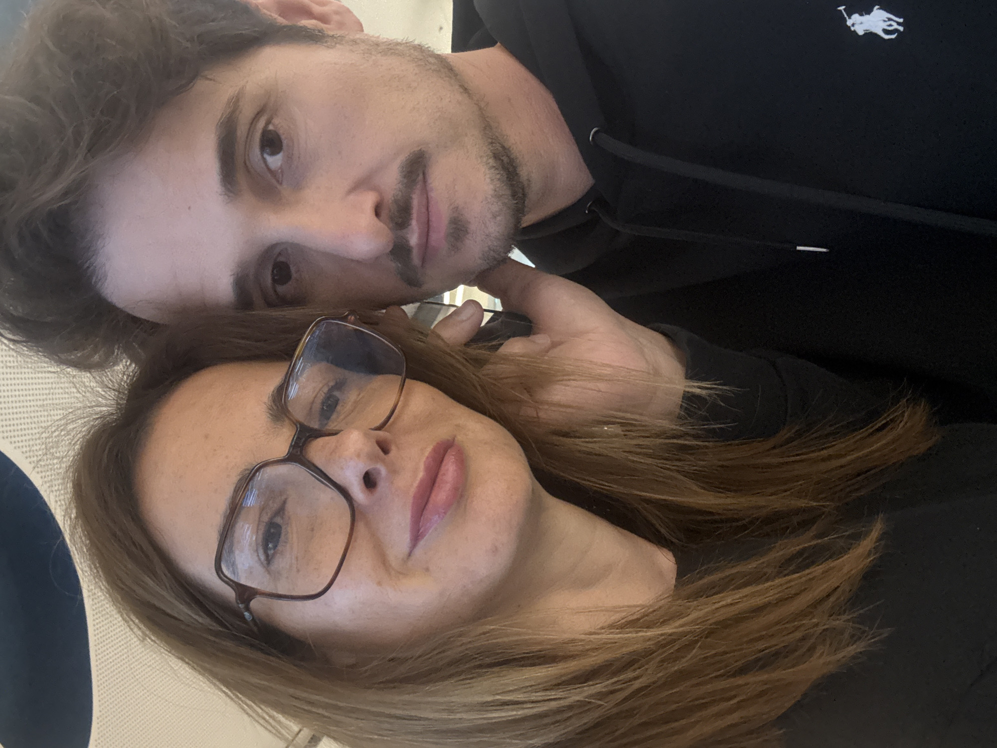 A selfie shot looking down at a man and woman, both wearing dark clothing. The man has dark hair and a mustache, and is wearing a black hoodie with a white polo player logo visible on the chest. The woman is wearing glasses and has light brown hair. The background is out of focus but appears to be the interior of a room, possibly an airplane based on a blue and white textured seat in the left side of the image. No information suggests this photo was taken in Lisbon, Portugal.