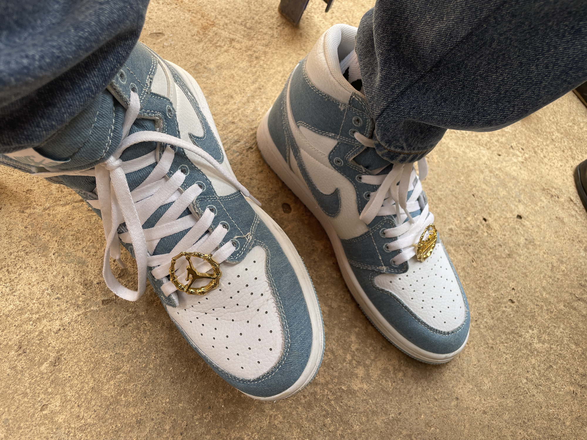 The image shows a pair of light blue and white high-top sneakers being worn. The shoes have white laces and a gold-colored charm with a figure on each lace. The person wearing the sneakers is also wearing light blue denim jeans. The surface beneath the shoes is a textured, beige-colored material. There are no indications that this image was taken in Akwanga, Nigeria.