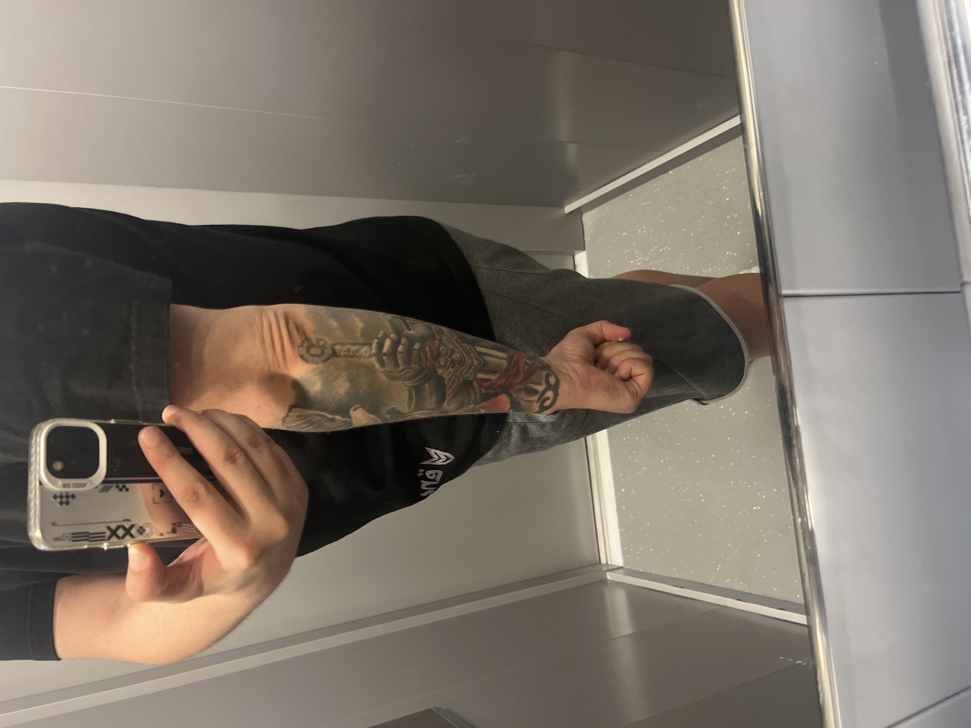 A person stands in what appears to be an elevator, holding a smartphone up to take a mirrored selfie. The person is wearing a black t-shirt and grey shorts. Their left arm, visible in the reflection, is covered in a tattoo depicting a knight's gauntlet holding a sword, with other elements below, possibly part of a larger design. The background shows the mirrored walls of the elevator and a speckled grey floor. The person's right arm is holding the phone, obscuring their face. The image quality is clear. The location is Varna, Bulgaria.