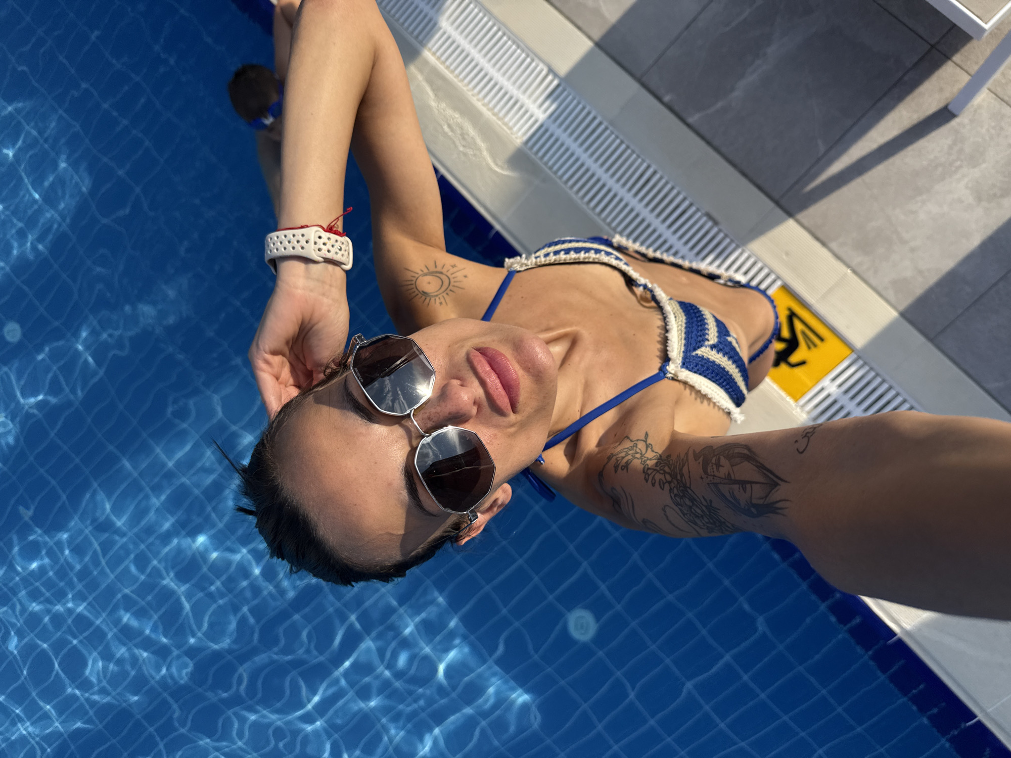 A woman in a blue and white crocheted bikini poses for a selfie at the edge of a swimming pool. She wears oversized sunglasses and a white smartwatch with a red string bracelet. Tattoos are visible on her left arm and shoulder. The pool water is a bright blue, and the surrounding pool deck is tiled with gray and white. In the background, another person is partially visible in the water. The scene appears to be at a hotel or resort in Aksu, Turkey, given the context.