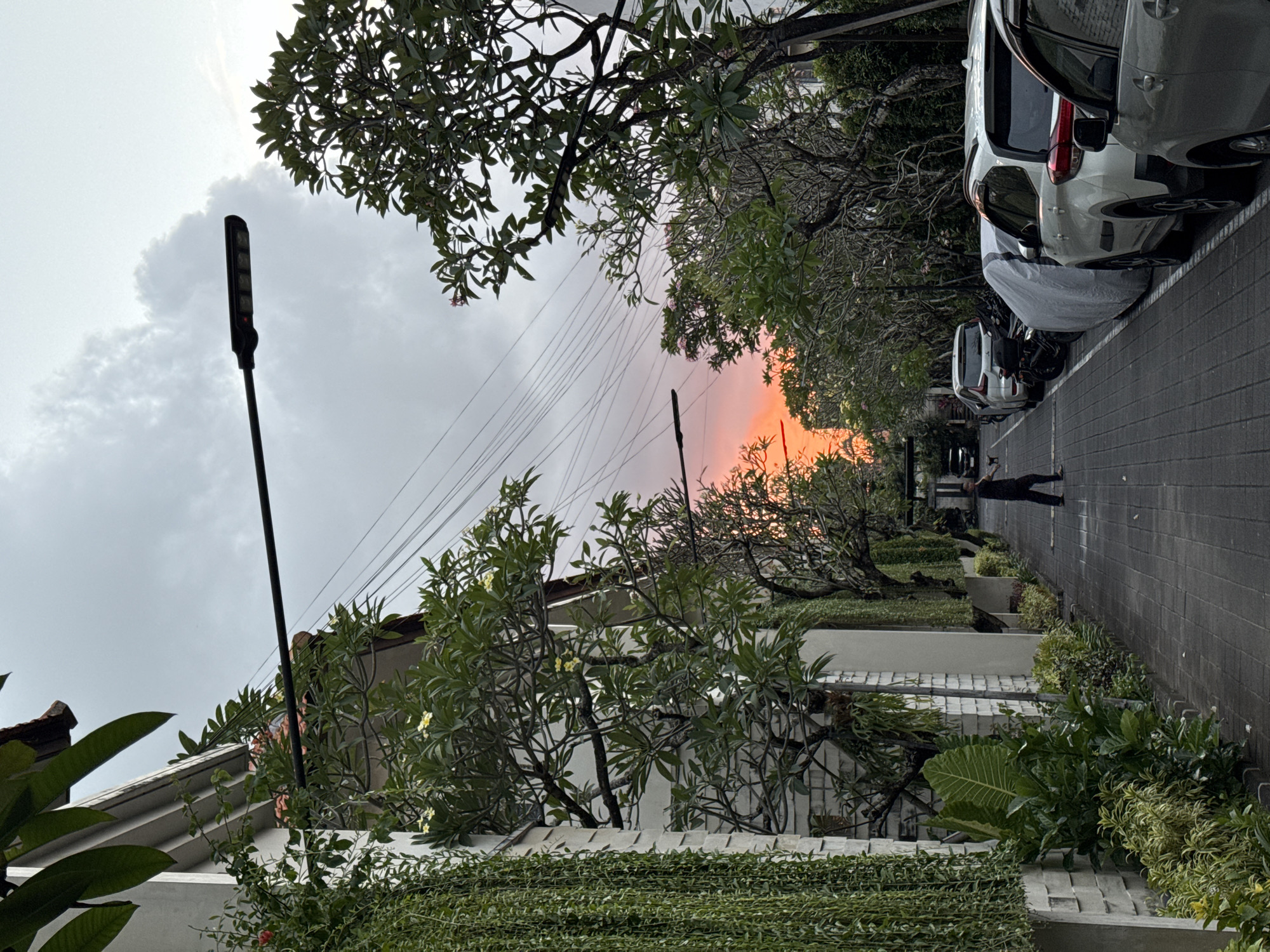 The image depicts a street scene in Denpasar, Indonesia, during twilight. A person walks down a paved walkway lined with parked cars and lush vegetation. Overhead, a street lamp is visible, and a bright orange sunset is peeking through the trees in the distance. The walkway is made of gray interlocking bricks, and a white wall with hanging plants runs along the left side of the street. Cars are parked on the right side of the street, with some covered by tarps. The sky is overcast with gray clouds, but the sunset casts a warm glow on the horizon. The overall atmosphere is calm and serene, suggesting a quiet evening in a residential or commercial area of the city.