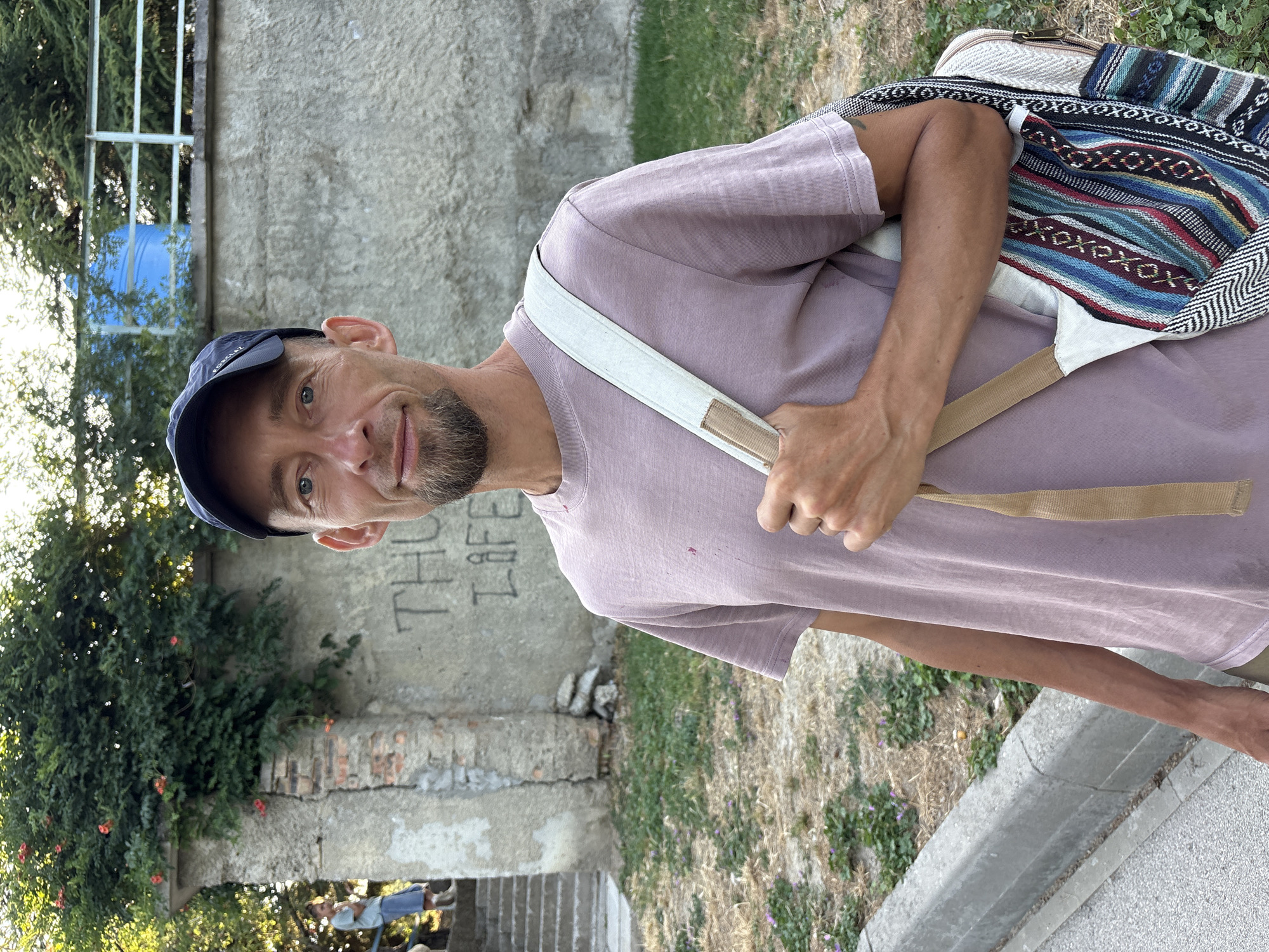 In Malyi Mayak, Ukraine, a fair-skinned man with a goatee and a blue baseball cap is shown from the chest up, standing outdoors. He is wearing a dusty rose t-shirt and carrying a bag with a colorful, patterned front over his right shoulder. The background features a textured concrete wall with graffiti, a stone structure with some greenery, and a blurred figure in the distance on a staircase. The ground is covered with dry grass and some small purple flowers, with a concrete curb and sidewalk in the foreground.