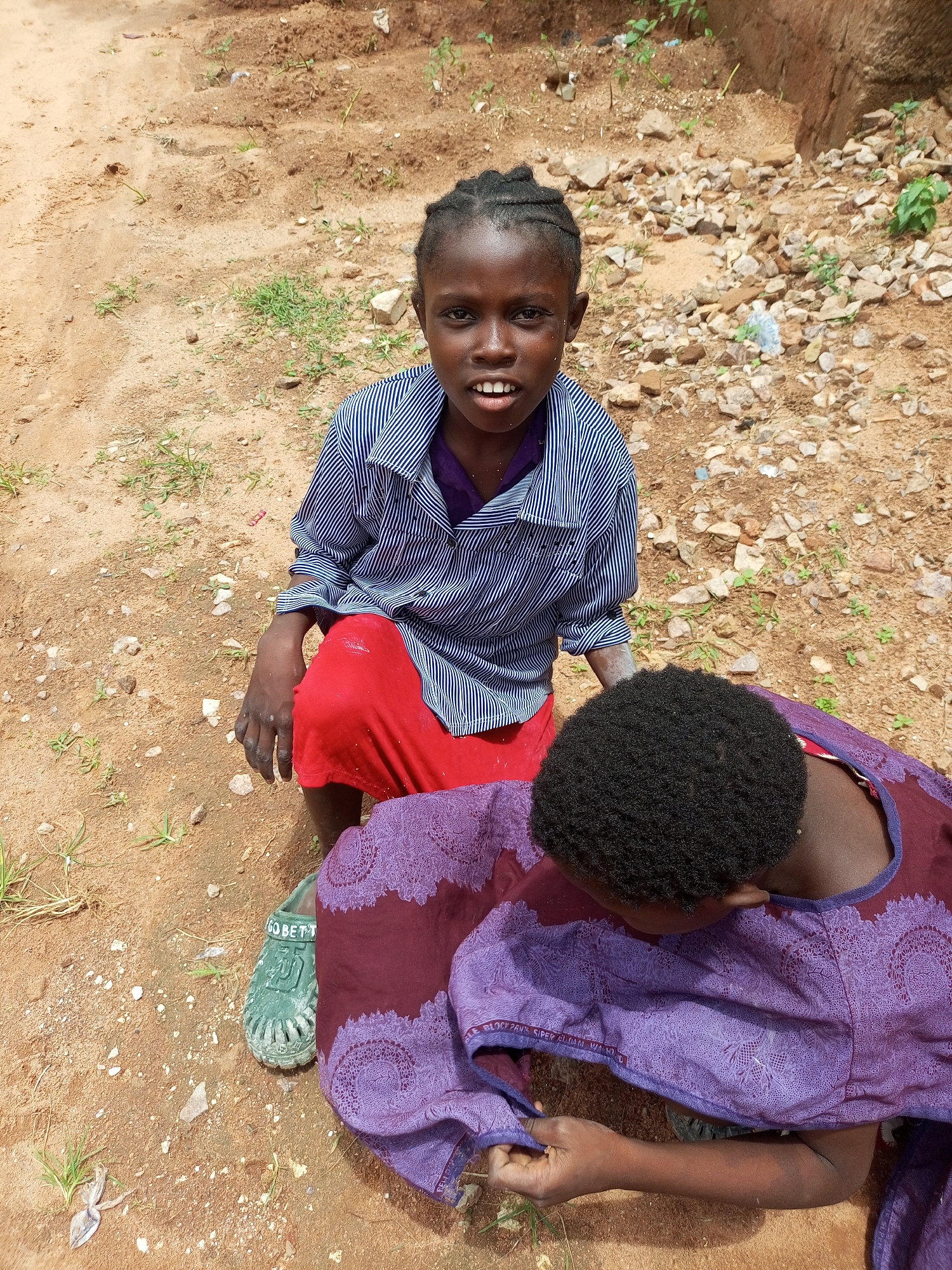 Two children, one kneeling and one in front of them, are in an outdoor environment with dirt and rocks on the ground. The kneeling child, wearing a blue and white striped shirt and a red skirt, is looking towards the camera with their mouth open. They have braided hair. The second child, whose back is to the camera, is wearing a purple and maroon patterned garment and has dark, curly hair. A green shoe with writing on it is visible near the kneeling child's foot. The ground is a mixture of sand and small stones with some sparse green vegetation. The scene appears to be in an informal, possibly rural, setting in Tempure, Nigeria.