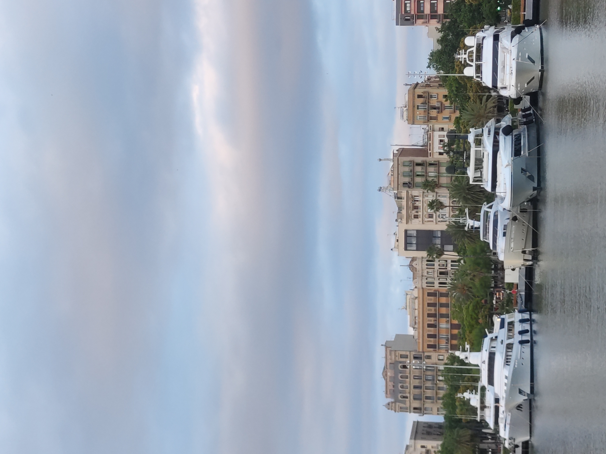The image captures a waterfront scene in Barcelona, Spain, likely in the Port Vell area. A row of large white yachts is docked in the foreground. The yachts are of varying sizes and designs, with prominent superstructures, windows, and railings. The water is calm, reflecting the sky and the boats.
Behind the yachts, a line of multi-story buildings forms the city's skyline. These buildings exhibit a mix of architectural styles, with some featuring ornate facades and balconies, while others are more modern in appearance. Lush green trees, including palm trees, are interspersed between the buildings and along the waterfront, adding a touch of nature to the urban landscape.
The sky above is partly cloudy, with soft, diffused light suggesting it might be early morning or late afternoon. The overall atmosphere is serene and peaceful, showcasing a blend of maritime activity and urban architecture characteristic of Barcelona's coastal districts. No people are visible in the image.
