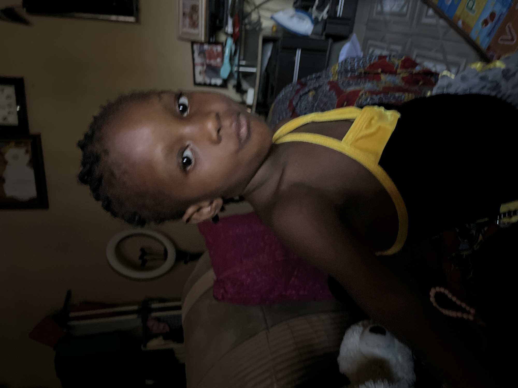 A child with dark skin and short, braided hair is lying down, wearing a black and yellow sleeveless top. They are looking directly at the camera with a neutral expression. The background includes a wall with framed pictures, a ring light, and furniture. A pink pillow and a white stuffed animal are near the child. There is no information within the image to confirm the location. The description doesn't contain any information about Madalla, Nigeria.