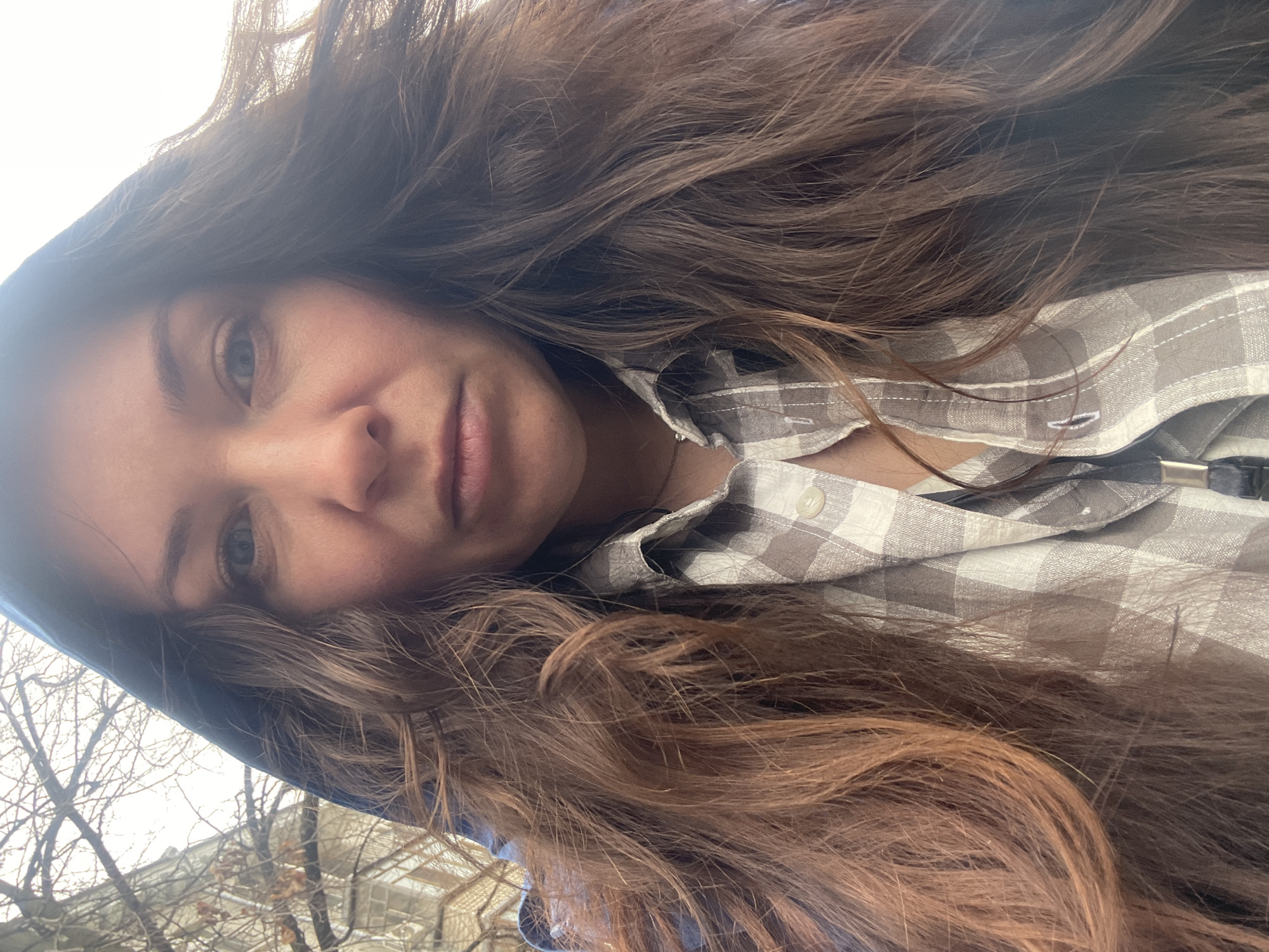 A person with long, wavy brown hair and fair skin is wearing a button-down plaid shirt. The person is wearing a blue hooded jacket. The background shows bare tree branches and a light-colored building. The environment suggests an outdoor location. Based on the image content, it is not possible to confirm the specific location in the city of Kazan, Russia.