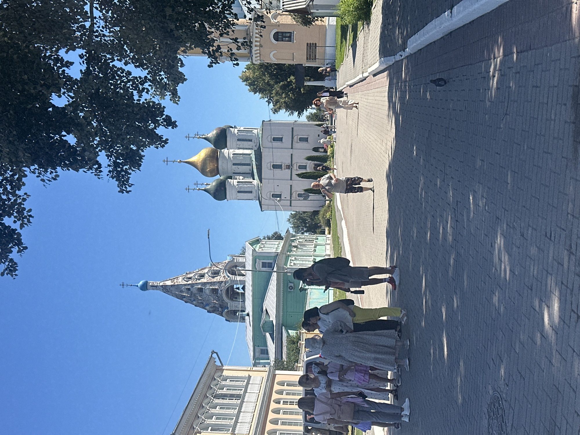 In Kolomna, Russia, on a bright, sunny day, people are gathered on a cobblestone street lined with trees. To the left, a group of tourists, including women and children, are conversing. In the background, an ornate green building with a tall, pointed bell tower with a blue dome and a cross stands prominently against a clear blue sky. Further down the street, a large, white cathedral with golden domes and crosses is visible, alongside another yellow building with a tower. Several individuals are walking along the street, including a man in the foreground running and a woman in a light-colored dress walking away from the camera. A pigeon is seen on the street. The overall scene depicts a lively tourist area with notable historical and religious architecture.
