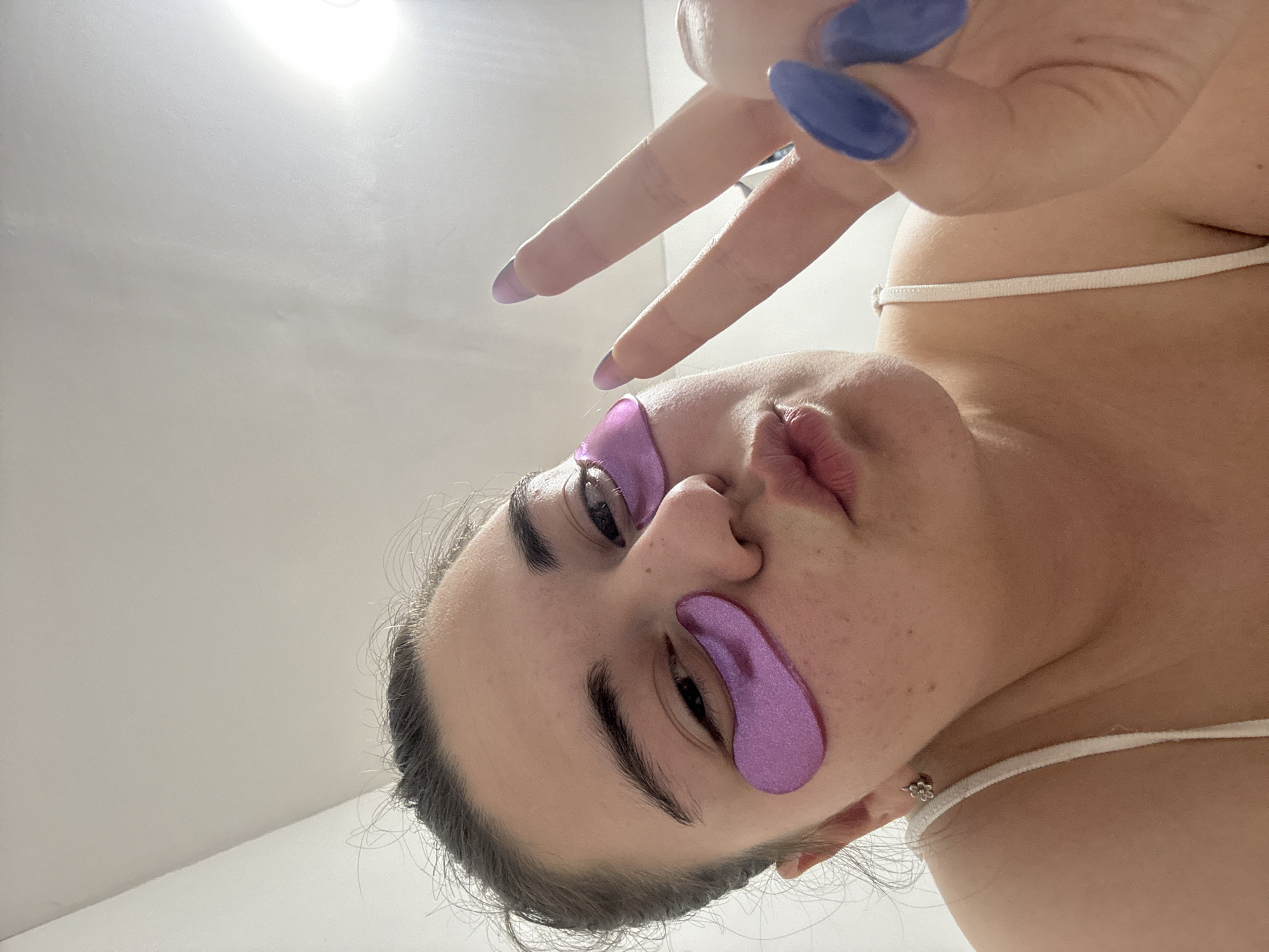 A woman with light skin is positioned in the lower right half of the frame, facing upward with her head tilted to the left. She has dark hair pulled back and is wearing purple under-eye patches. Her nails are painted a dark blue-purple color, and she is making a peace sign with her fingers. The background is a plain white wall. There is no indication of Zlatitsa, Bulgaria.
