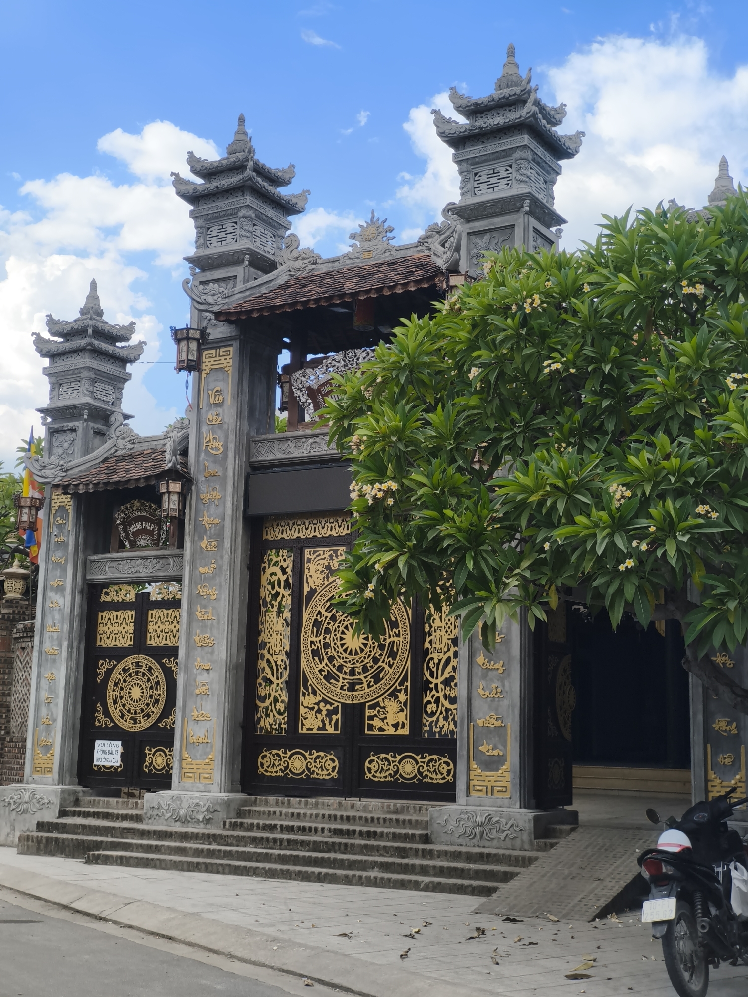 This is a medium shot, eye-level, vertical photograph of a temple gate in Nha Trang, Vietnam. The gate is elaborate, with gray stone pillars adorned with gold lettering and intricate designs. Large, dark double doors with ornate gold circular patterns stand in the center. Above the doors, a tiled roof is visible, supported by decorative stone elements. Flanking the gate are two tall, multi-tiered stone towers, also decorated with intricate carvings.
A lush green tree with small white flowers partially obscures the right side of the gate. The sky above is a bright blue with scattered white clouds, suggesting it is daytime. In the foreground, concrete steps lead up to the gate, and a ramp is visible to the right. A black and white motorcycle is parked near the ramp.
The text visible on the gate includes Vietnamese characters and a sign that reads "VUI LÒNG KHÔNG ĐẦU XE TRƯỚC CỨNG TAM QUAN," which translates to "Please do not park your vehicle in front of the Tam Quan gate." The overall impression is one of grandeur and cultural richness.