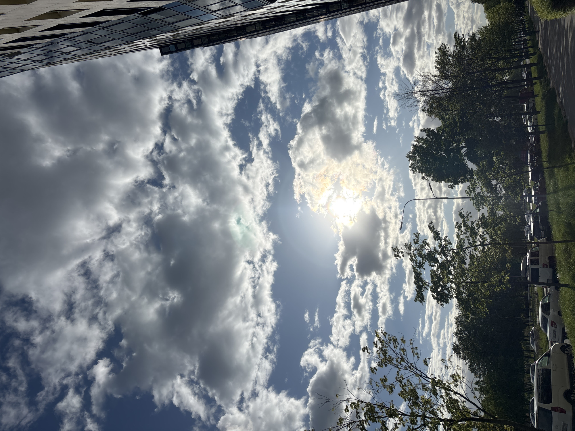 The image shows a view looking up towards the sky in an urban environment. In the top left corner, a portion of a modern building with reflective glass windows is visible. The sky is blue with scattered white clouds, and the sun shines through a gap in the clouds, creating a bright glow. On the right side of the image, trees with green leaves line a street or path. Parked cars are visible along the street.