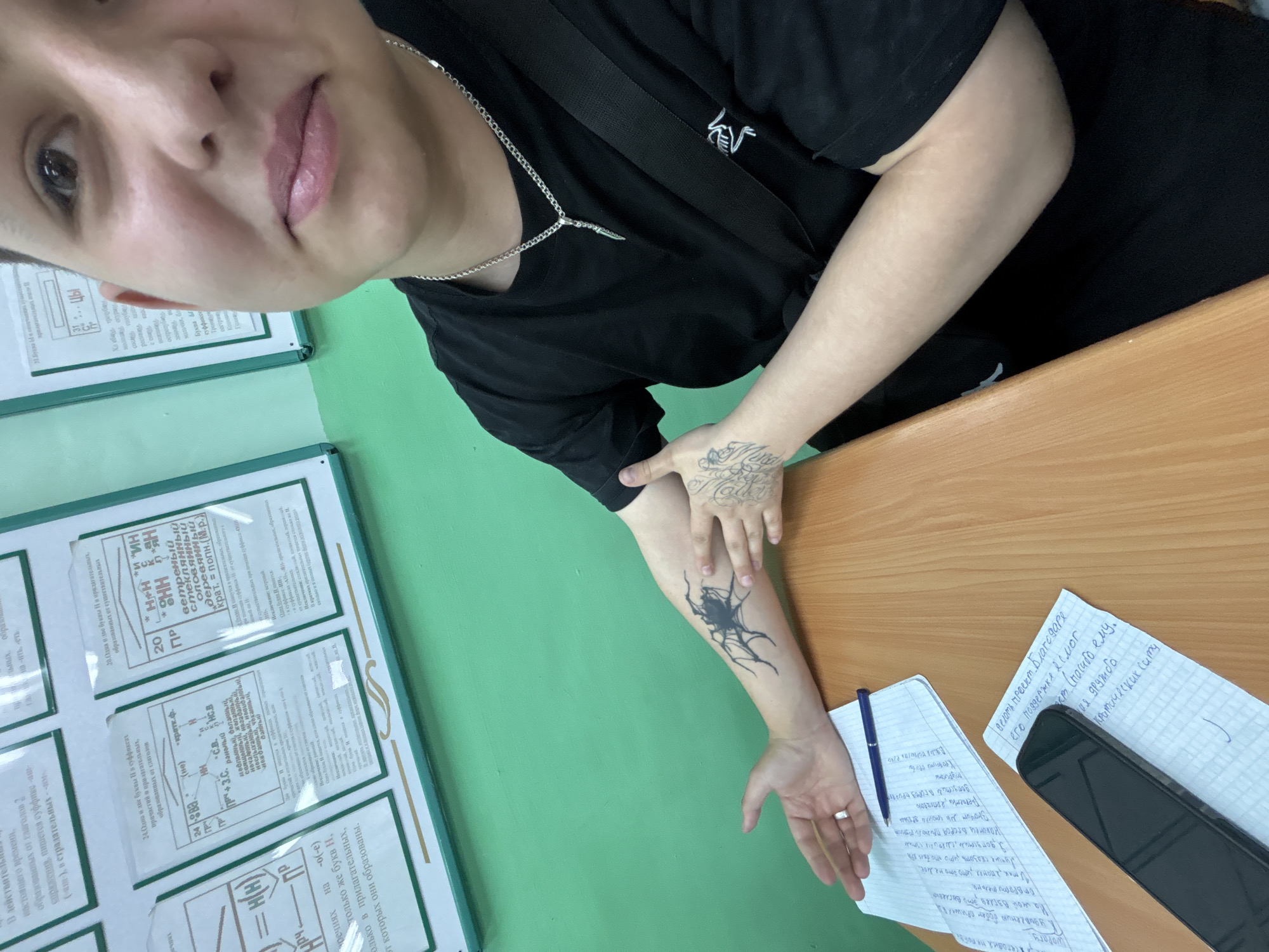 The image captures a selfie in what appears to be a classroom or study environment in Birobidzhan, Russia. The main subject is a person, likely female, with a slight smile, looking directly at the camera. They are wearing a black t-shirt and a silver chain necklace with a dagger-like pendant.
Notable details on the person include prominent tattoos on their right arm and hand. One on the forearm is a stylized, abstract spider or insect-like design in black ink. Another on the back of the hand features intricate script or calligraphy. A ring is visible on their left hand's ring finger.
The background features a vibrant green wall with several white-framed charts containing text in Russian. These charts appear to be educational materials, likely related to grammar or linguistics, discussing rules for "Н" and "НН" in adjectives and participles, with examples such as "ветреный", "стеклянный", "оловянный", and "деревянный", and sections on "Исключения" (Exceptions) and "Краткие прилагательные" (Short adjectives).
On a light wooden desk in the foreground, there are two sheets of paper with handwritten notes in Russian, a blue pen, and a black smartphone. The handwritten notes include phrases like "делать проект Благодаря", "его поддержке есмог", "Спасибо ему", "кратических ситу", "Наконец вгород прилого резист", and "Отвратительне", alongside other personal thoughts or creative writing snippets. The overall scene suggests a moment captured during study or a break in a learning setting.